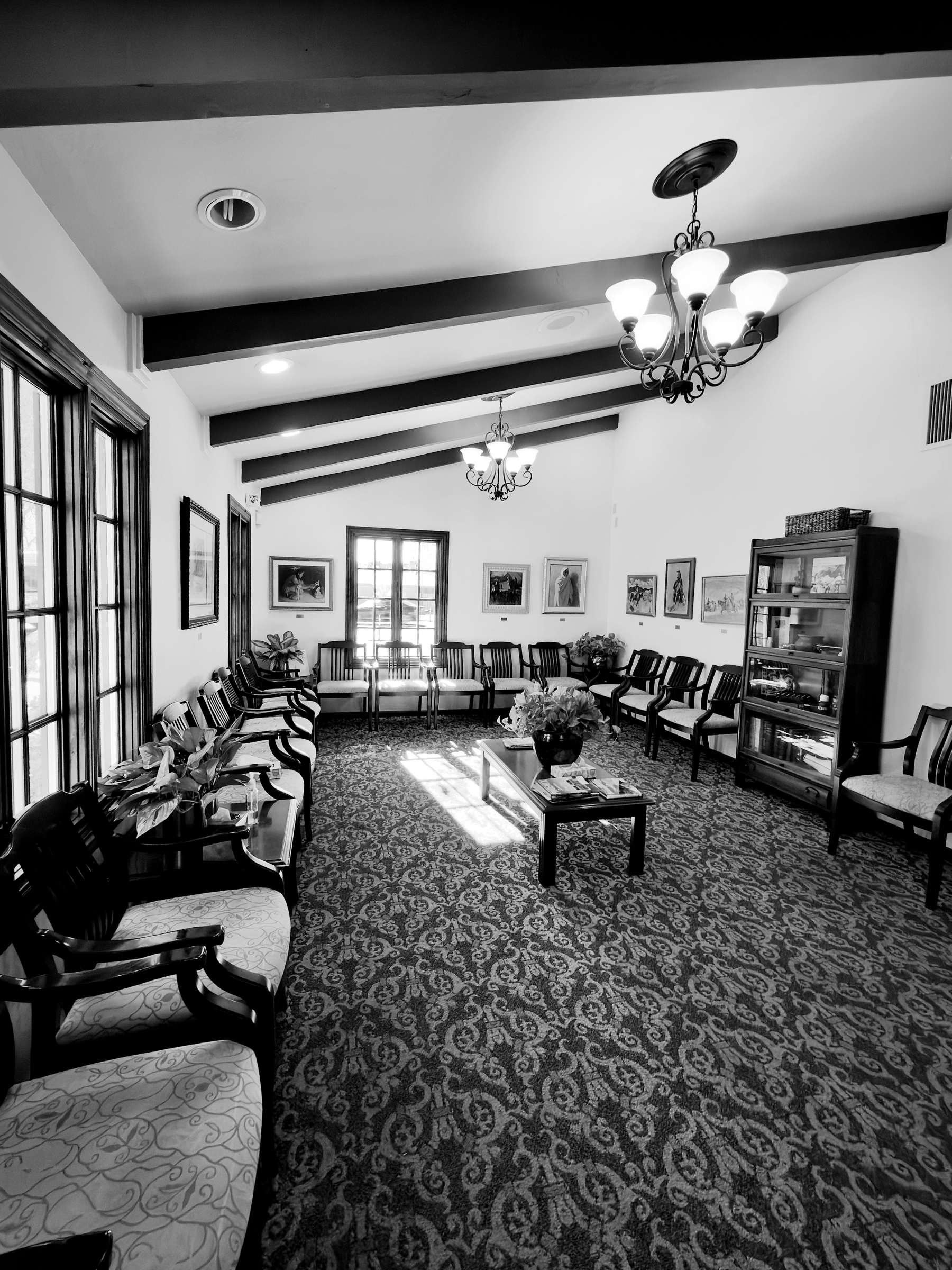 Auto-generated description: A spacious waiting room features rows of chairs along the walls, framed artwork, a patterned carpet, and elegant chandeliers hanging from a ceiling with exposed beams.