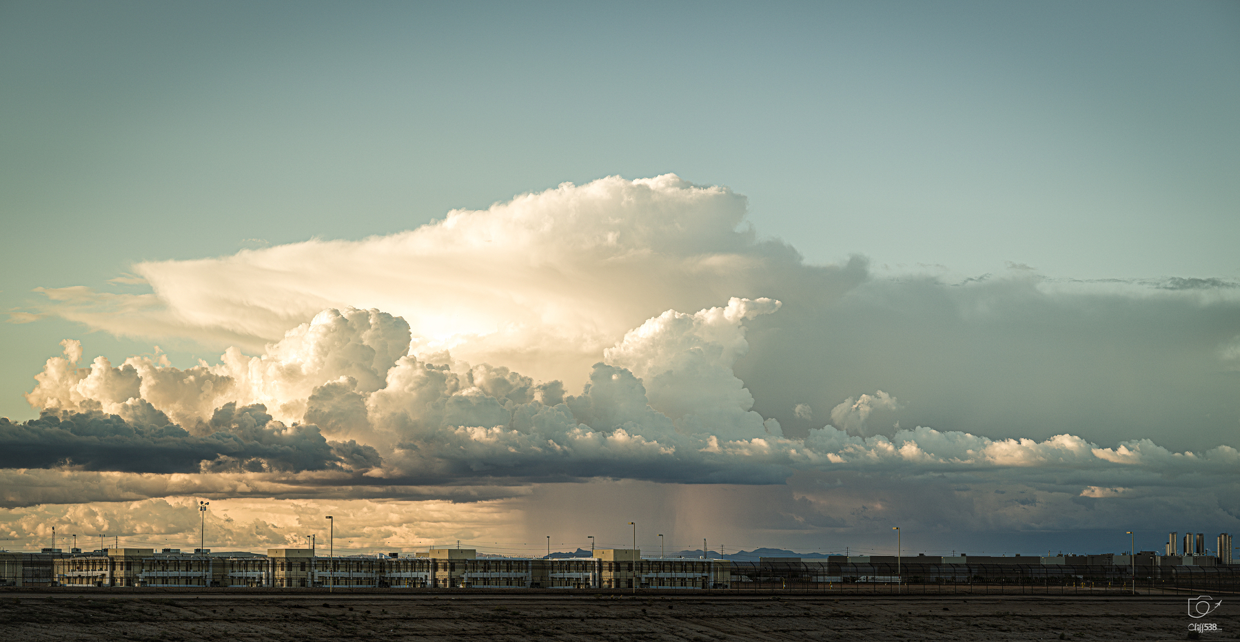 Auto-generated description: A dramatic sky filled with large, dense clouds hovering over a distant industrial or warehouse complex near a barren landscape.