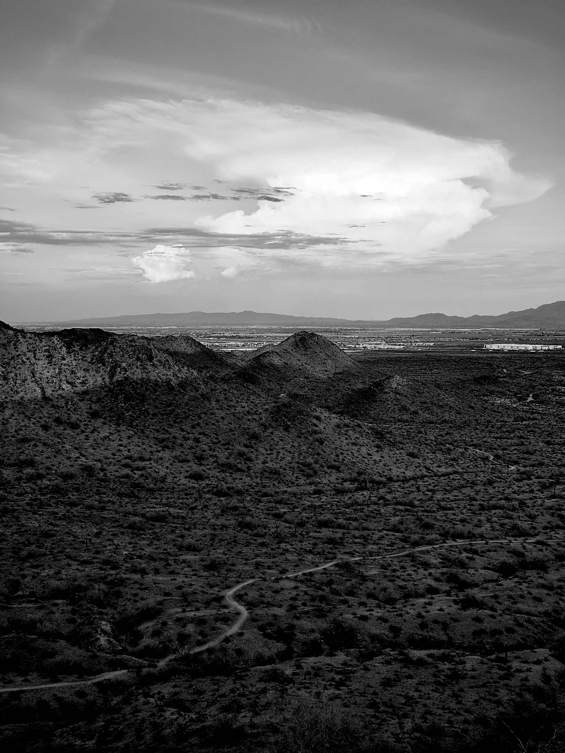 Auto-generated description: A winding trail cuts through a rugged desert landscape with hills in the midground and a vast sky filled with clouds above.