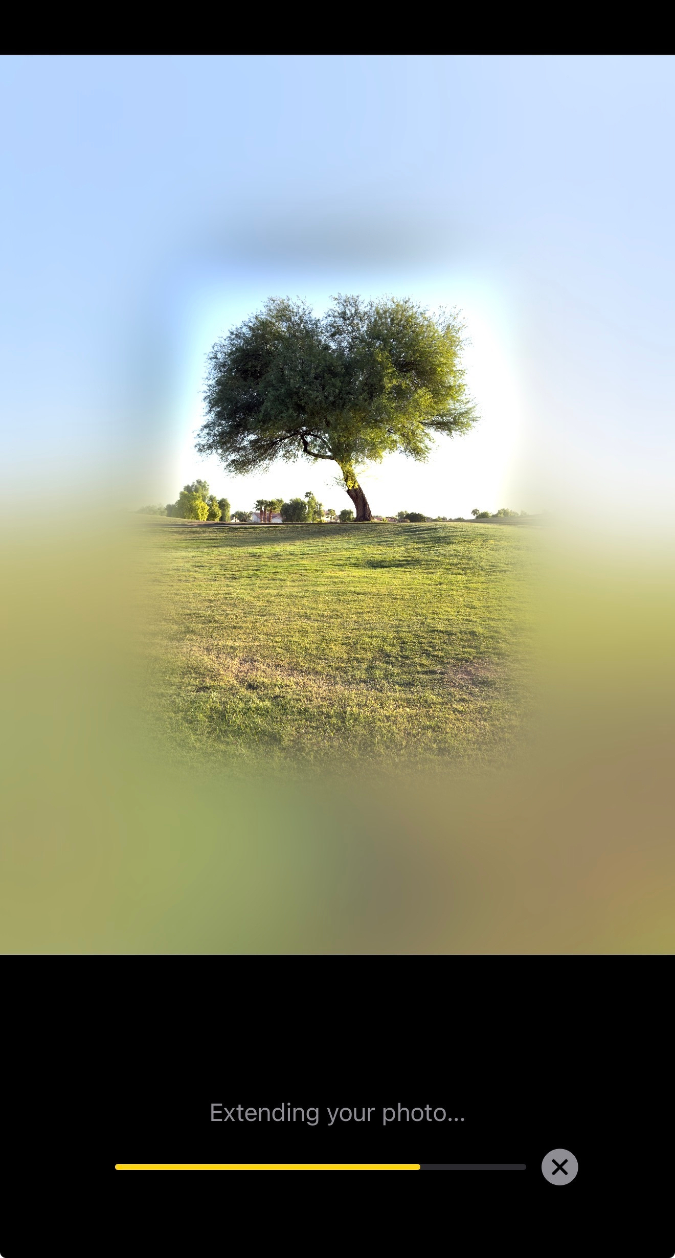 Auto-generated description: A solitary tree stands on a grassy field under a clear blue sky, with some distant trees and small structures in the background.
