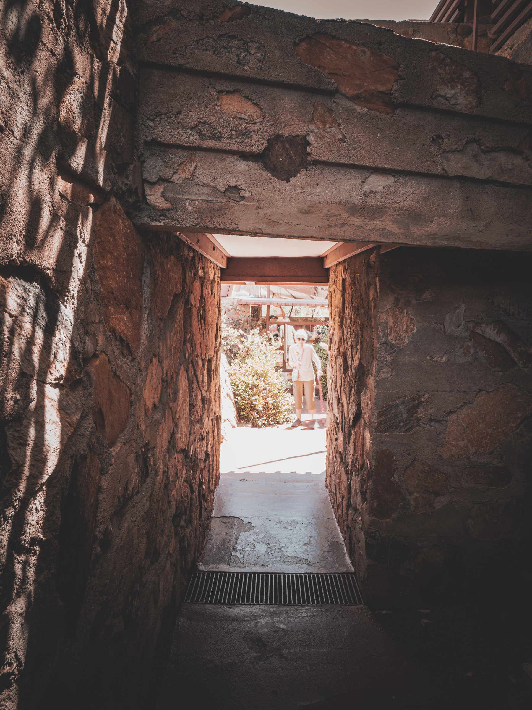 Auto-generated description: A stone passageway leads to a sunlit outdoor area with a person standing in the distance.