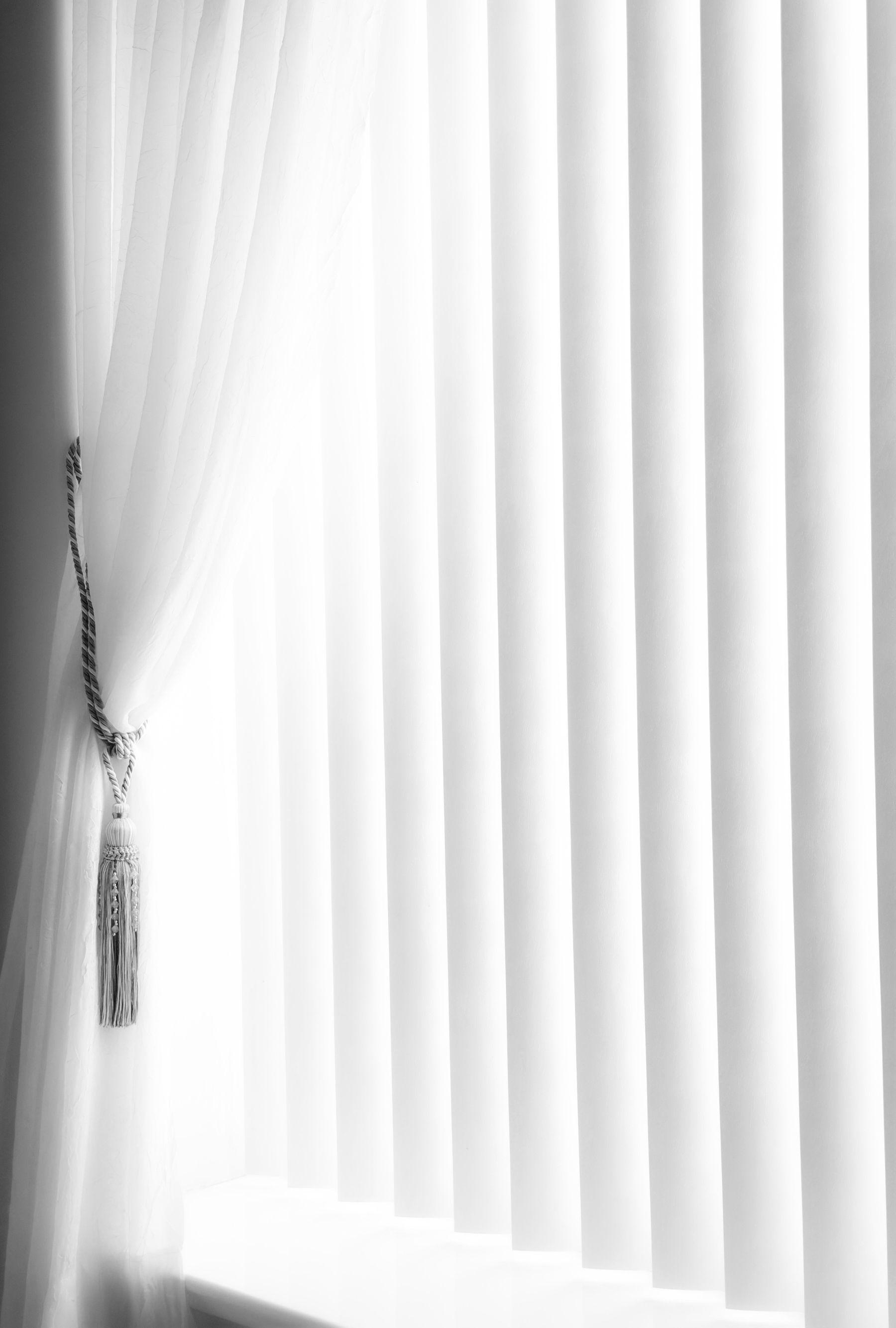 Auto-generated description: White vertical blinds cover a window, with sheer white curtains tied back with a decorative tassel on the left side.