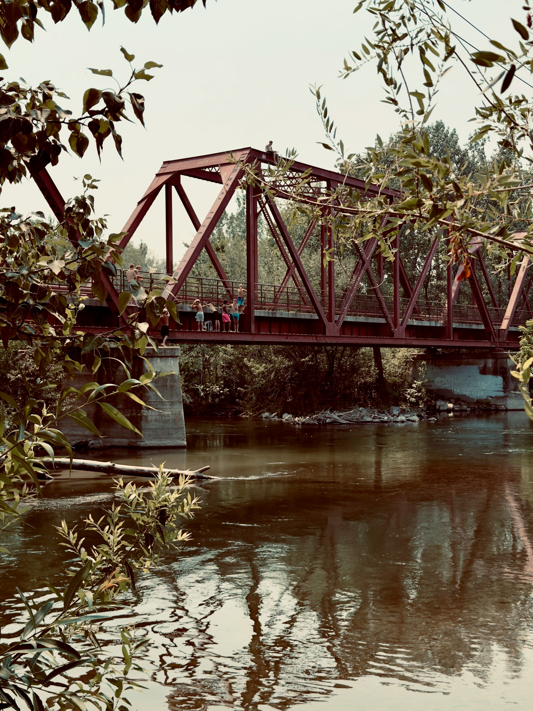 Auto-generated description: A red metal truss bridge spans over a calm river, surrounded by leafy trees, with a few people standing on the bridge railing.