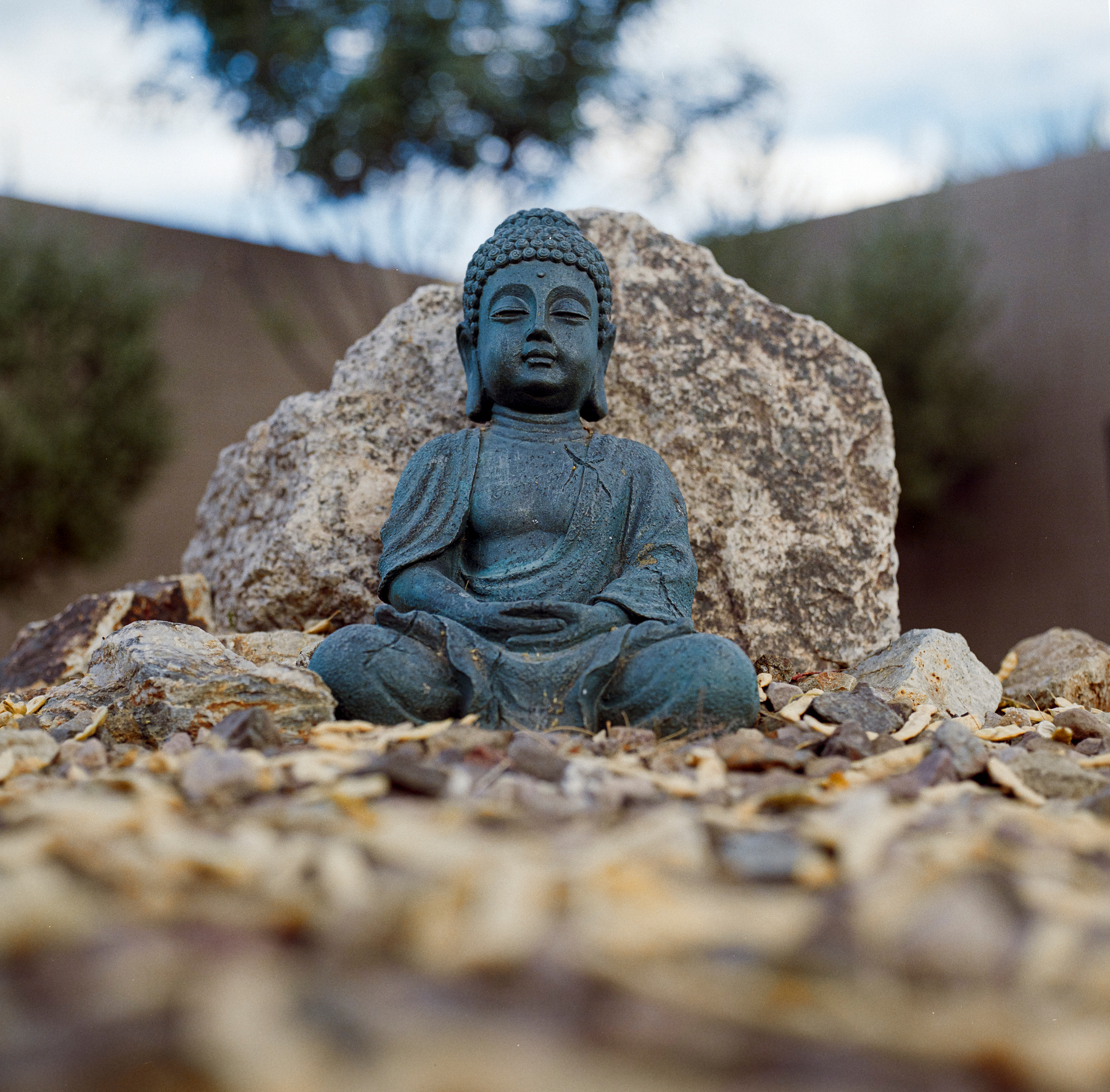 Auto-generated description: A small Buddha statue is seated in a meditative pose among rocks and pebbles with a large stone and blurred trees in the background.