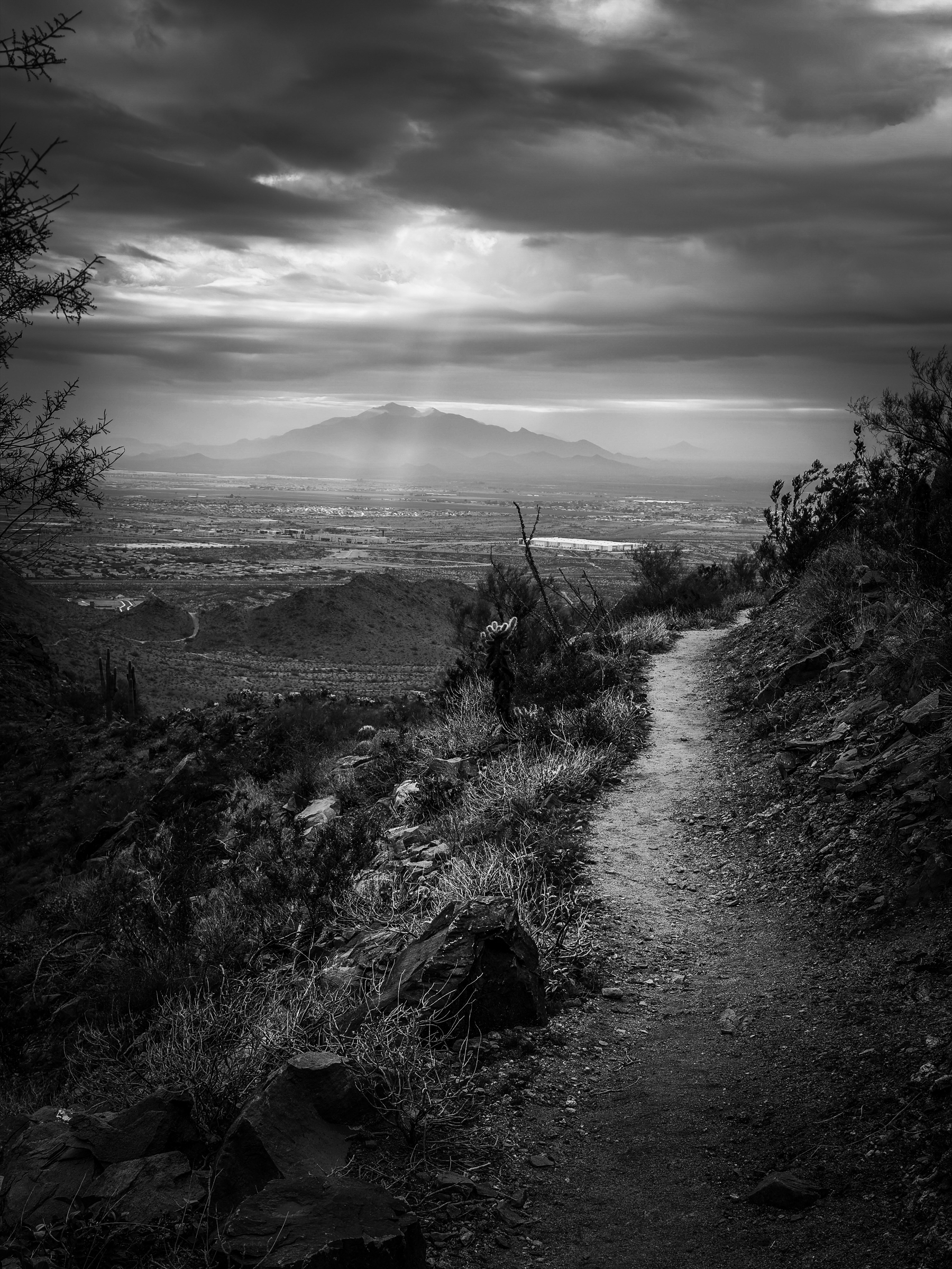 Auto-generated description: A narrow path winds through a rugged landscape under a dramatic cloudy sky, with distant mountains illuminated by sunrays in the background.