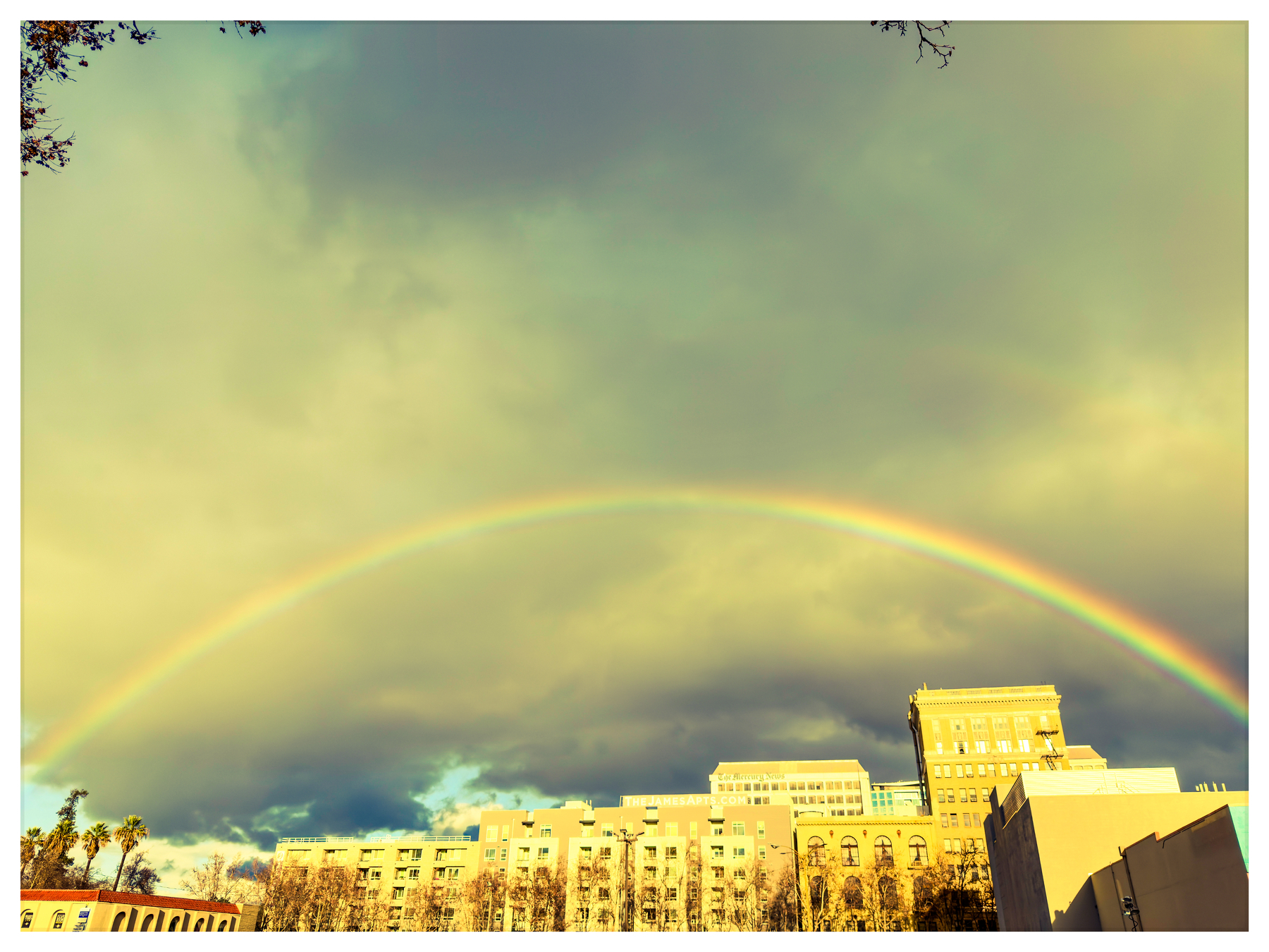 Auto-generated description: A vibrant rainbow arches over a cityscape with buildings under a cloudy sky.