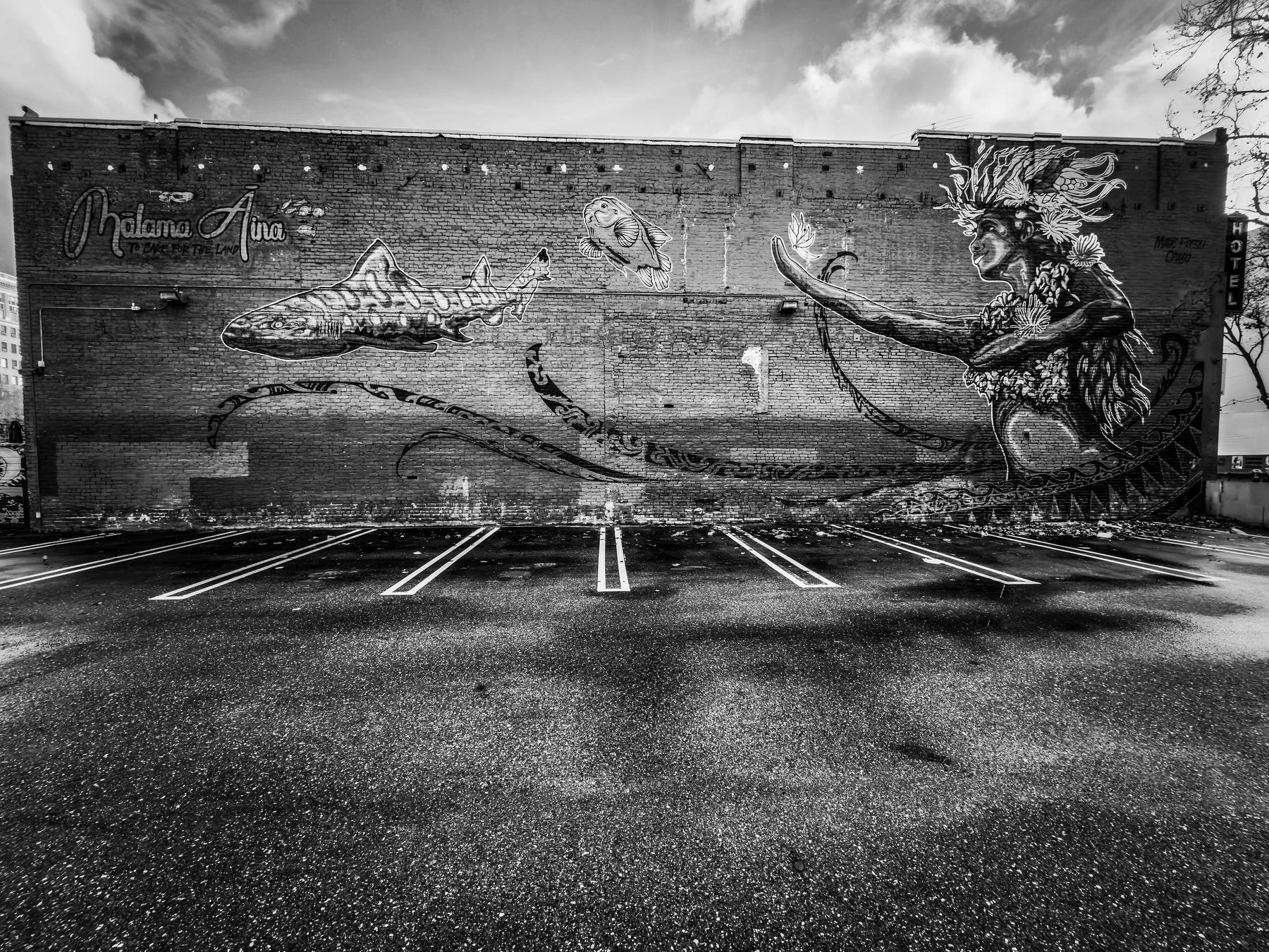 Auto-generated description: A black and white mural of a fantastical sea creature with a fish on a brick wall in an empty parking lot.