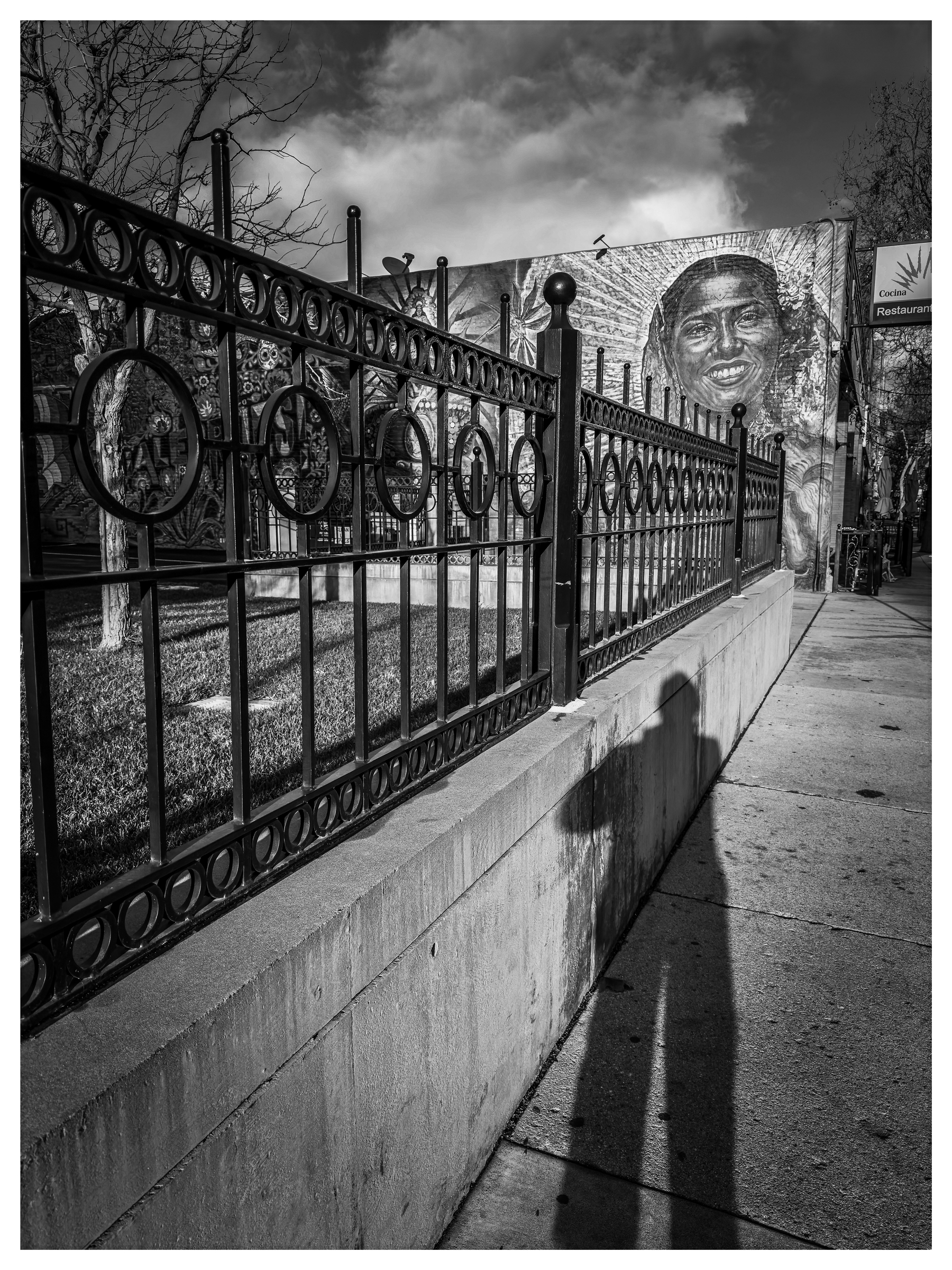 Auto-generated description: A shadow of a person with a hat is cast on a sidewalk next to an iron fence, with a mural of a face in the background.