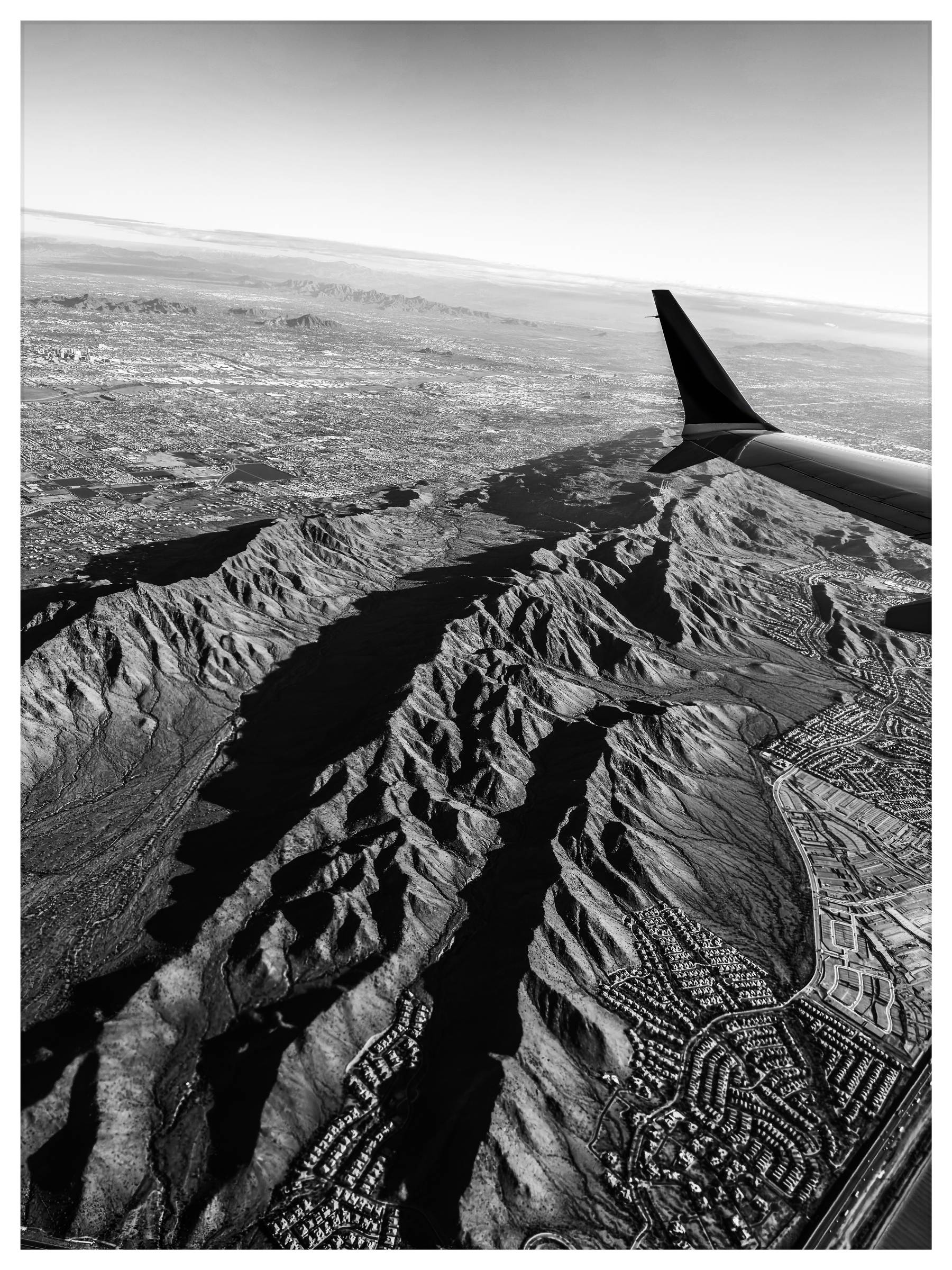Auto-generated description: Aerial view of rugged mountain terrain and a sprawling cityscape under an airplane wing, captured in black and white.
