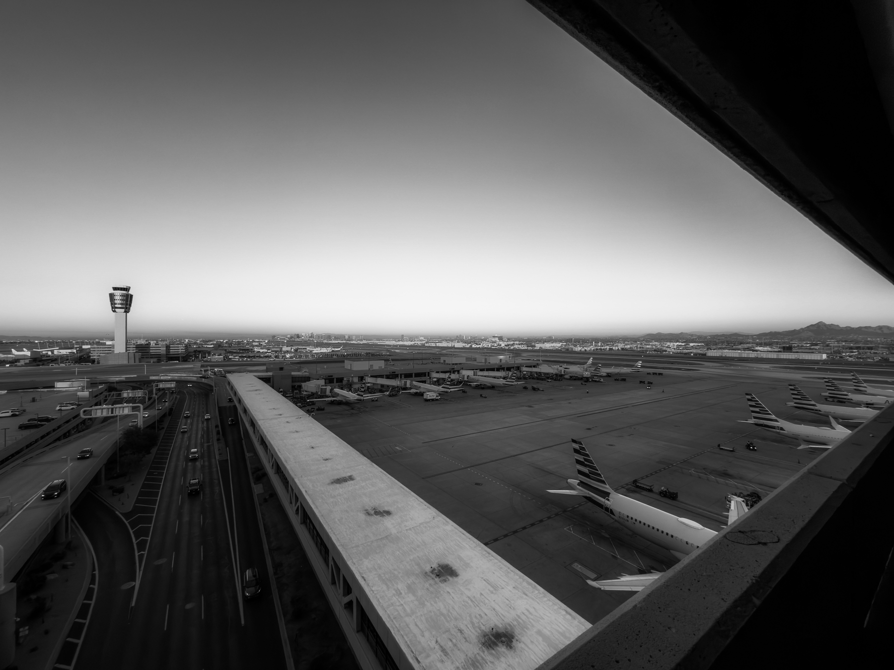 Auto-generated description: A black-and-white aerial view of an airport features planes on the tarmac, a control tower, and a highway running alongside.