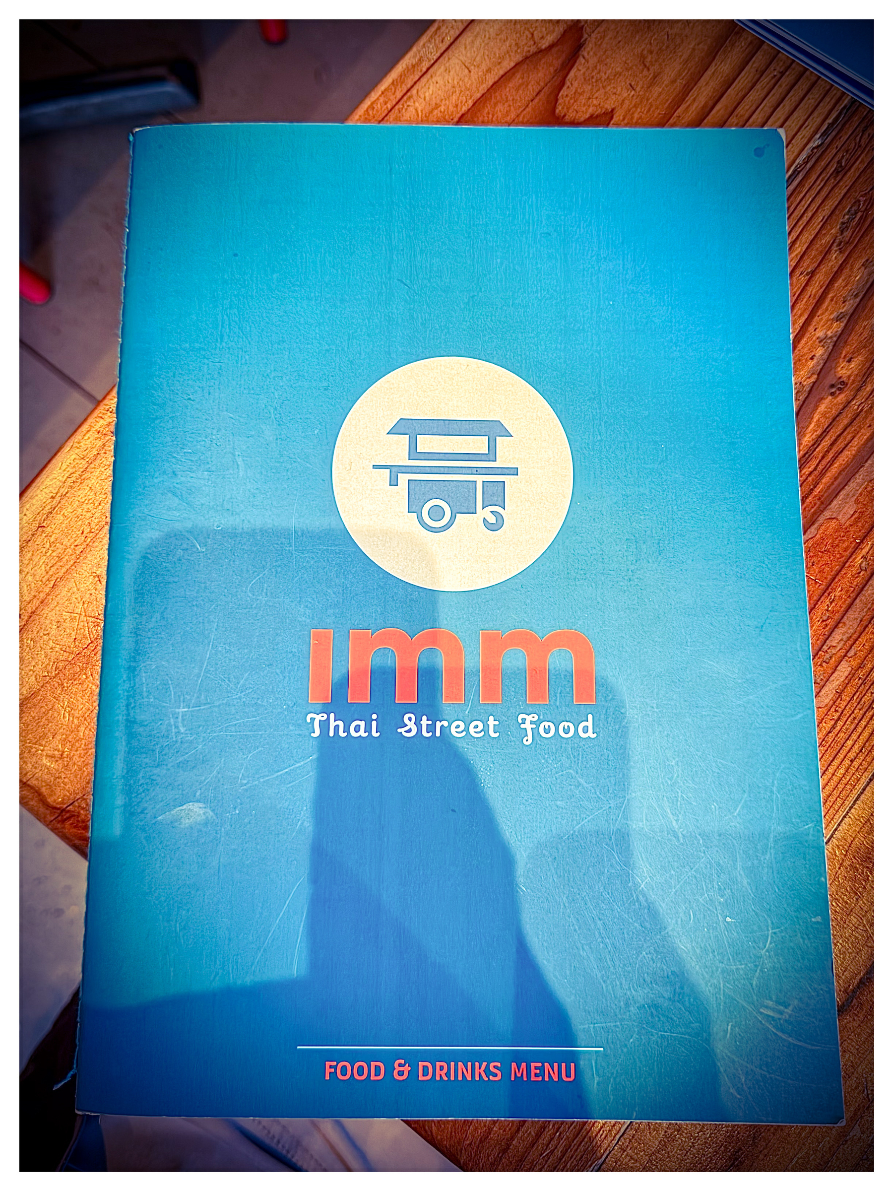 Auto-generated description: A blue menu cover with the logo and text imm Thai Street Food sits on a wooden table.