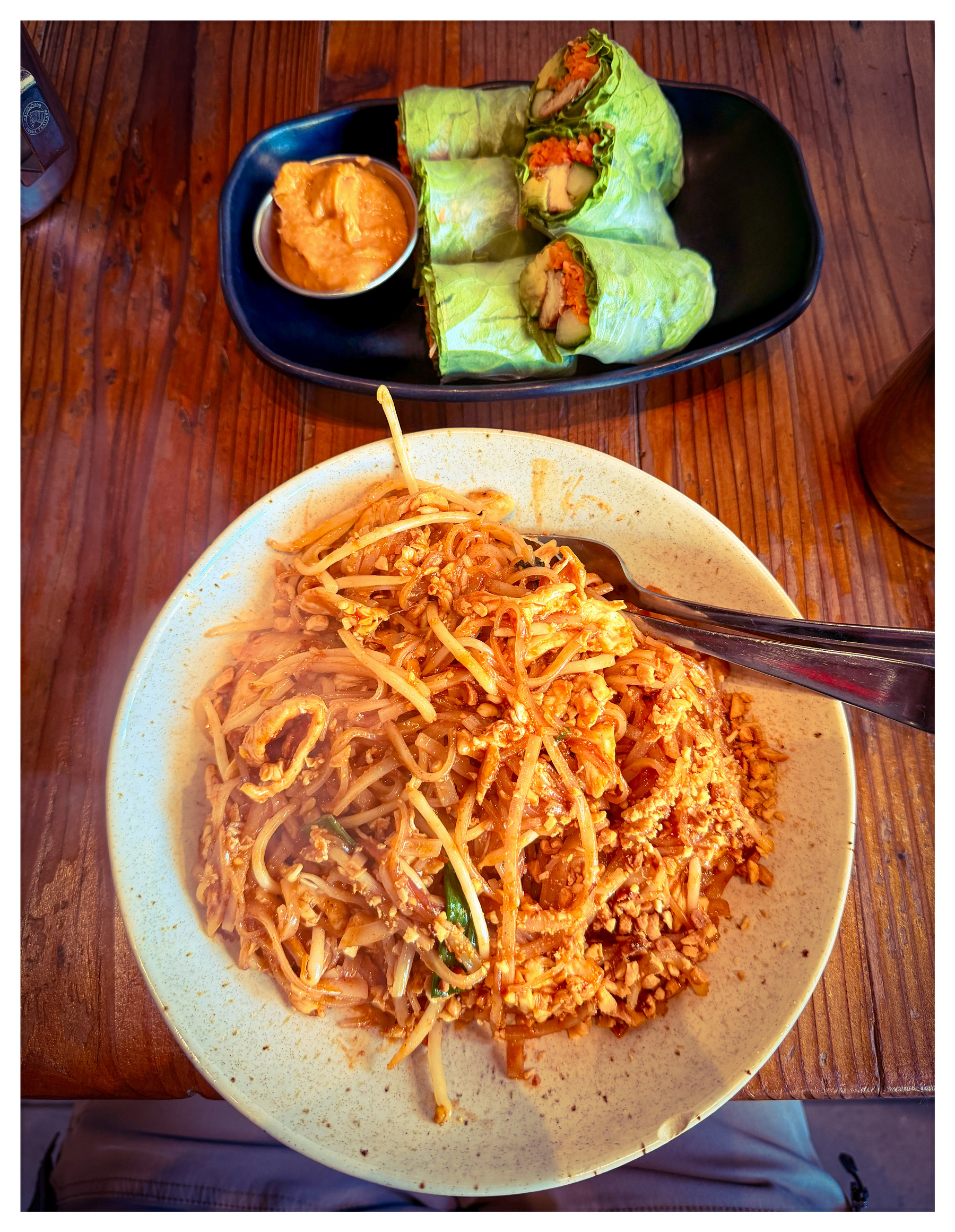 Auto-generated description: A plate of noodles with peanuts and vegetables is served alongside fresh spring rolls with a dipping sauce on a wooden table.