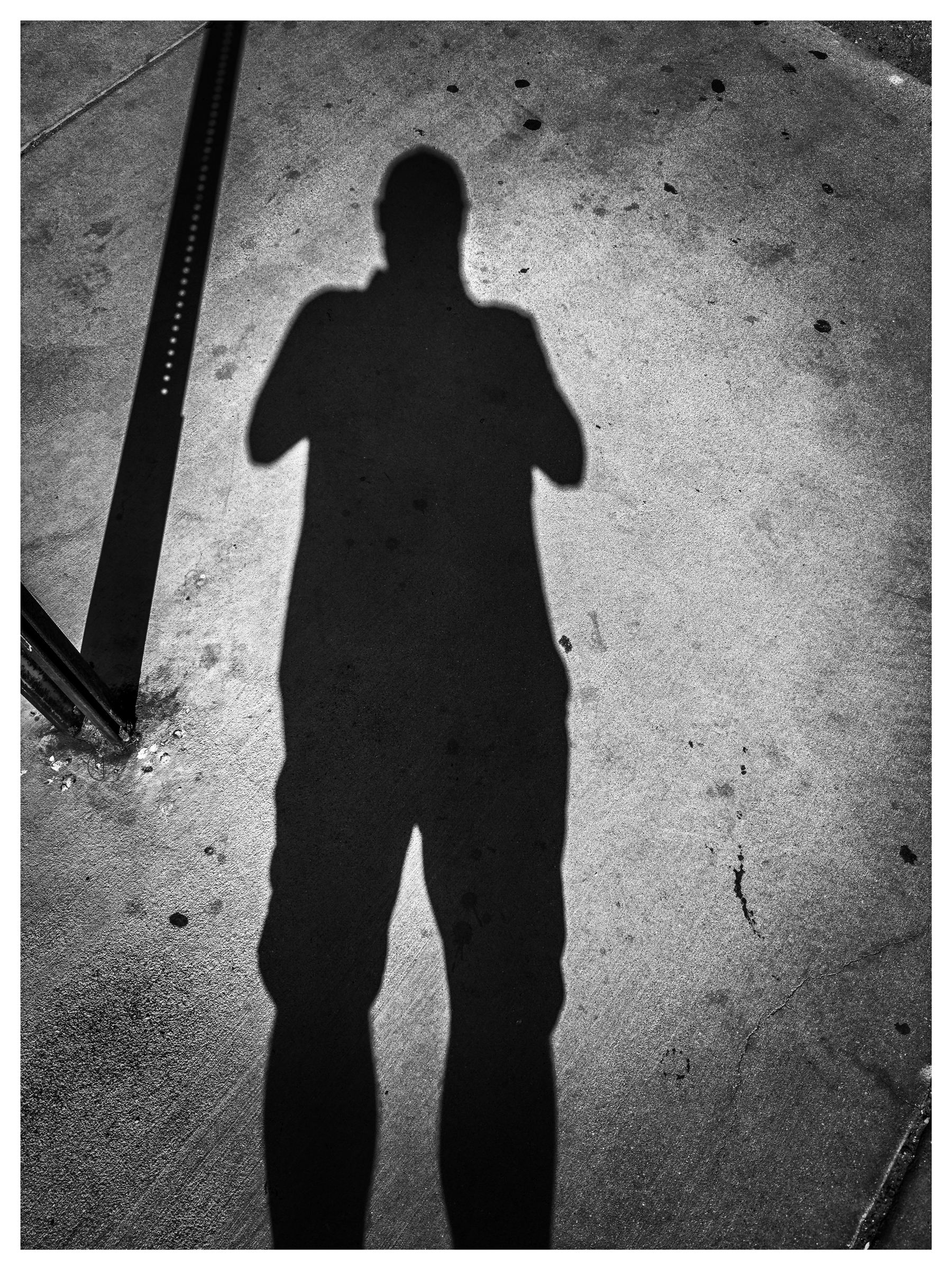 Auto-generated description: A shadow of a person taking a photo is cast on a paved surface, next to a vertical metal pole.