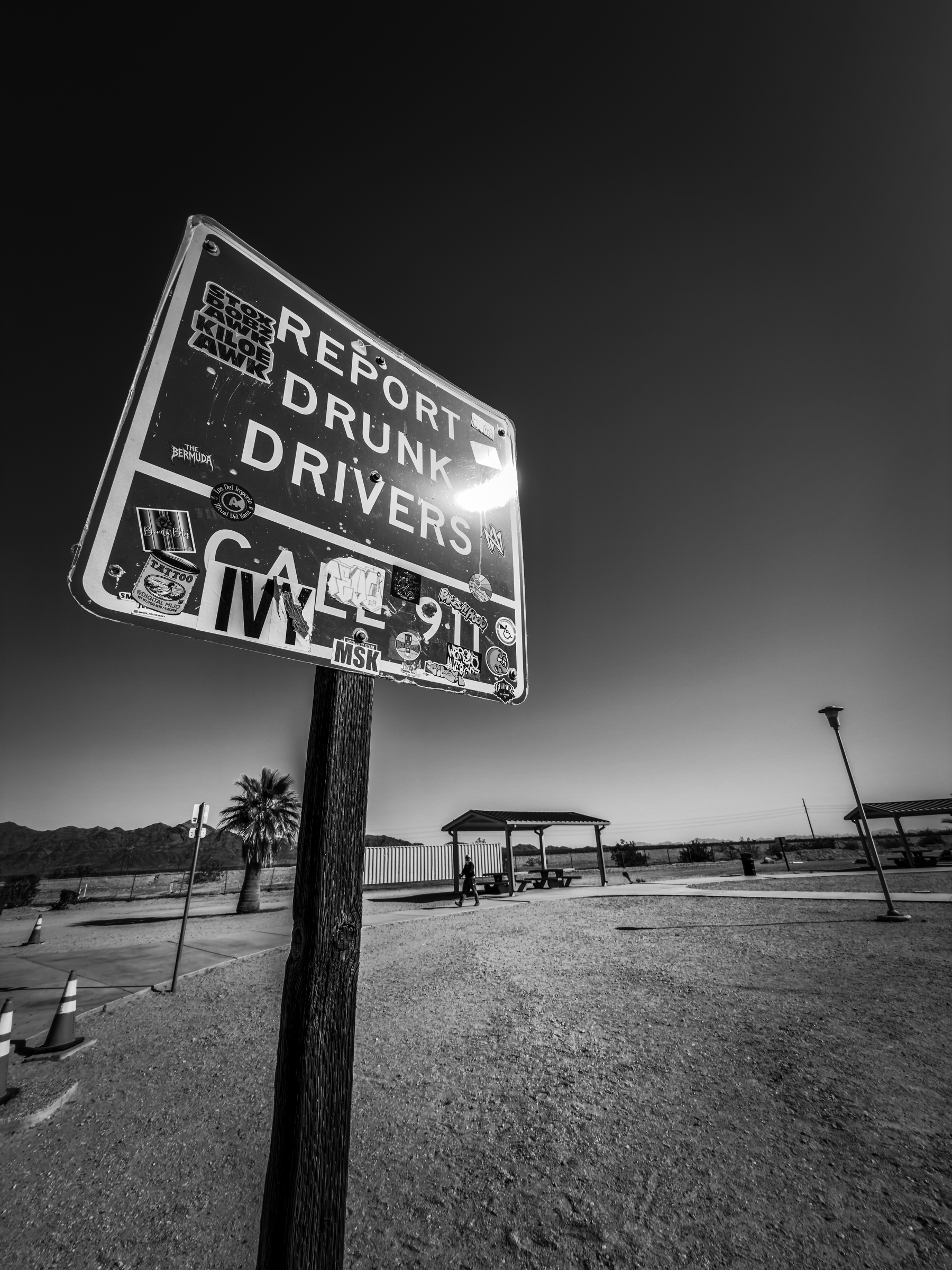 Auto-generated description: A sign urging the reporting of drunk drivers is covered with stickers and stands against a clear sky in a wide, open landscape with a distant view of a shelter and palm tree.