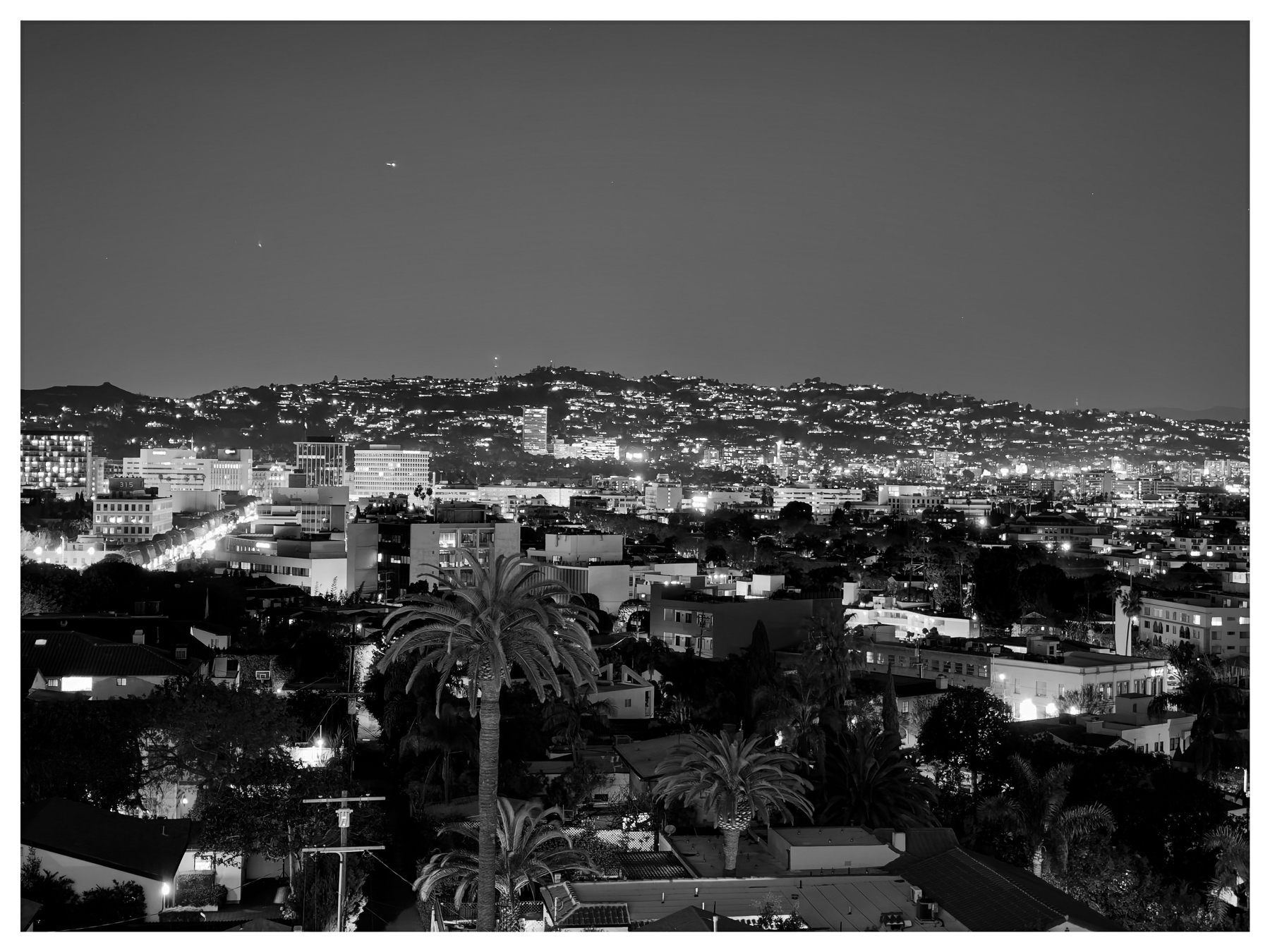 Auto-generated description: A cityscape at night features illuminated buildings with a backdrop of hills and a clear sky.