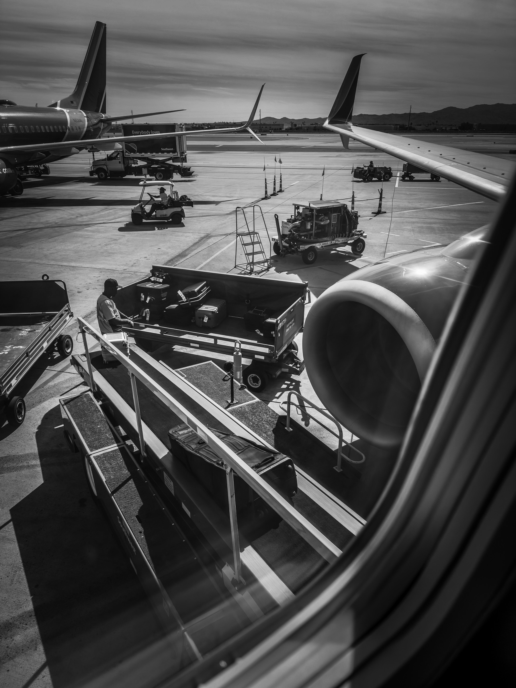 Auto-generated description: A black and white view from an airplane window shows airport ground crew handling luggage and carts near several parked airplanes.