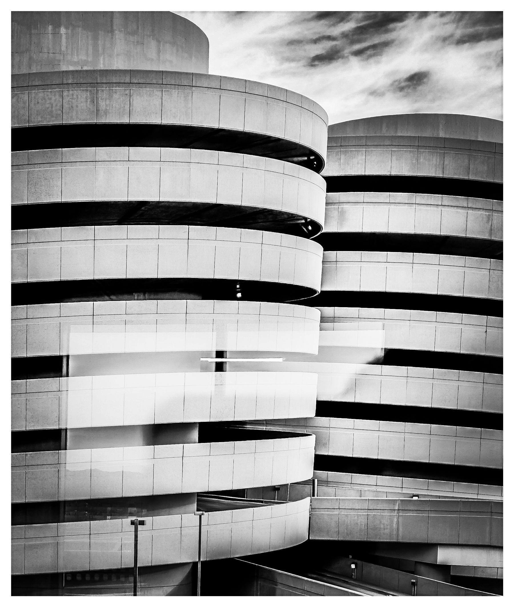 Auto-generated description: A black and white photograph captures a modern, cylindrical parking garage with spiral ramps and cloudy skies above.