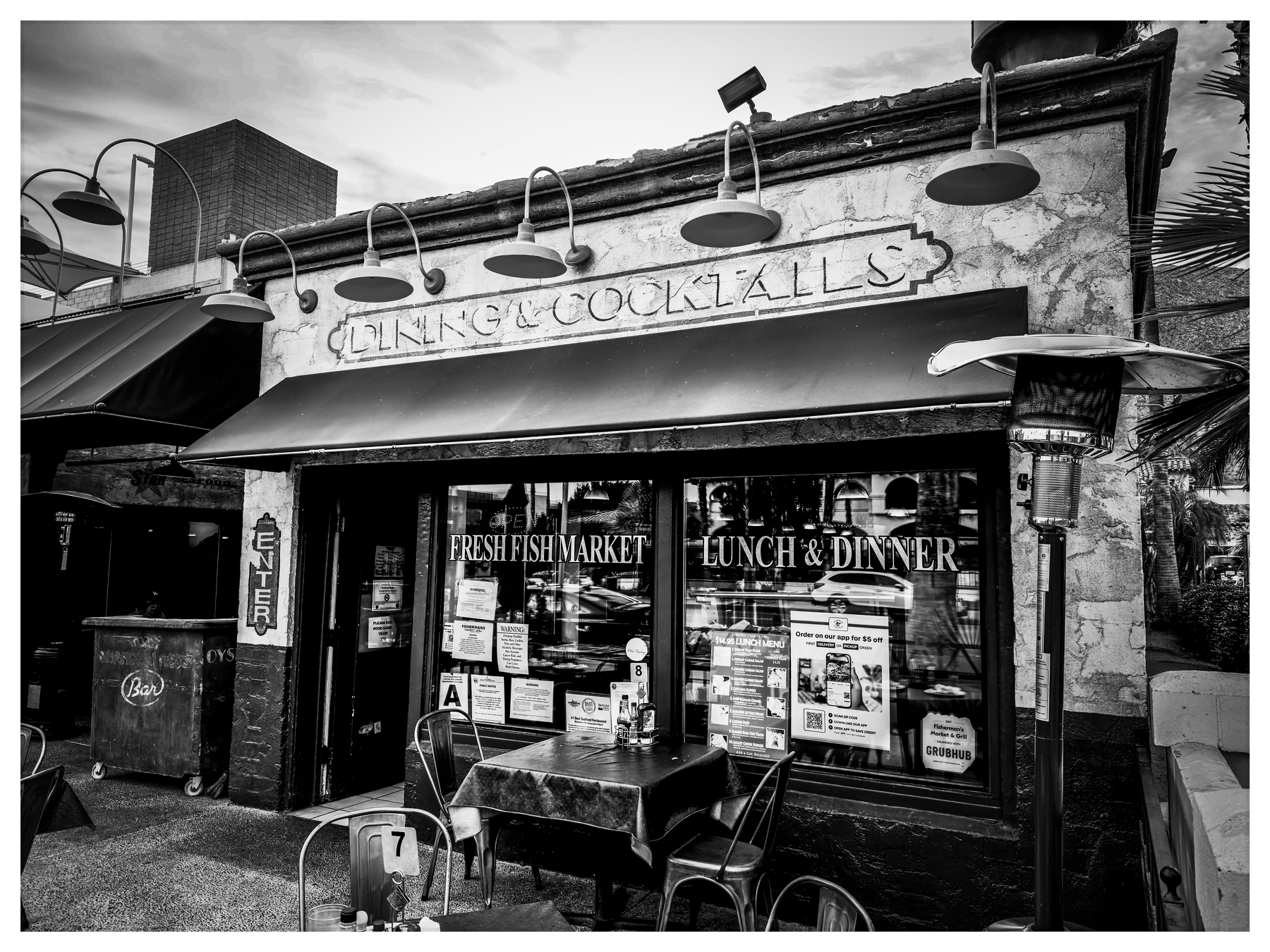Auto-generated description: A black and white photo of a rustic storefront with a sign for Drinks & Cocktails and Fresh Fish Market Lunch & Dinner, featuring outdoor seating and various posters in the windows.