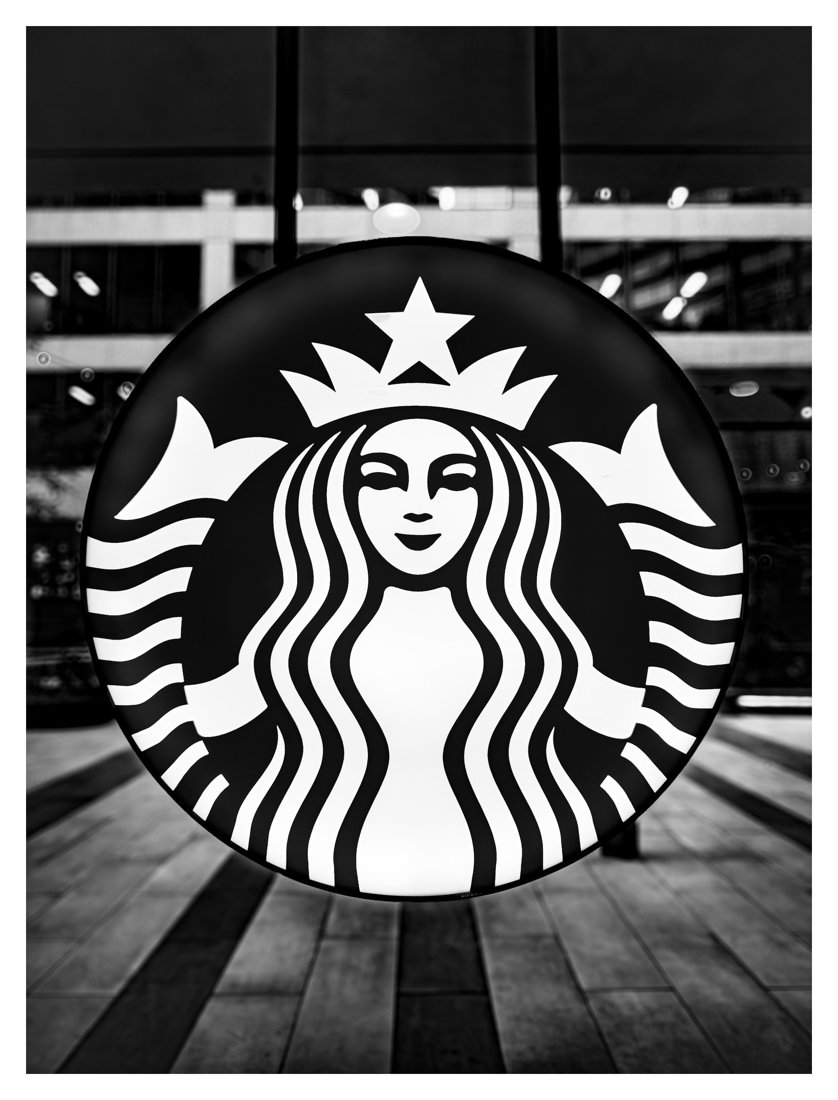 Auto-generated description: A black and white circular Starbucks logo featuring a crowned mermaid with flowing hair is prominently displayed against a background of glass windows and a tiled floor.