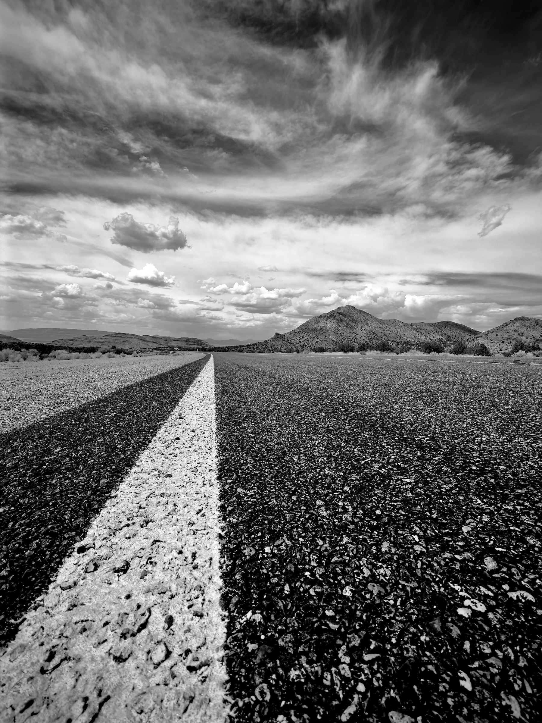 Auto-generated description: A long, empty road stretches toward distant mountains under a dramatic cloudy sky in black and white.