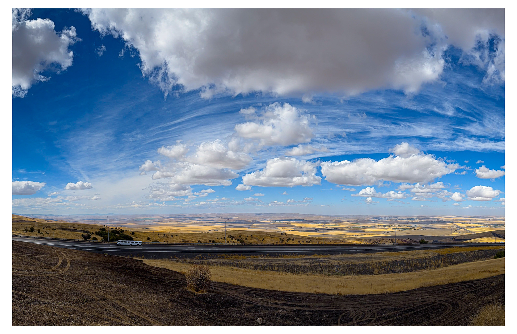 Auto-generated description: A vast landscape with rolling hills under a partly cloudy blue sky and a road running through the foreground.