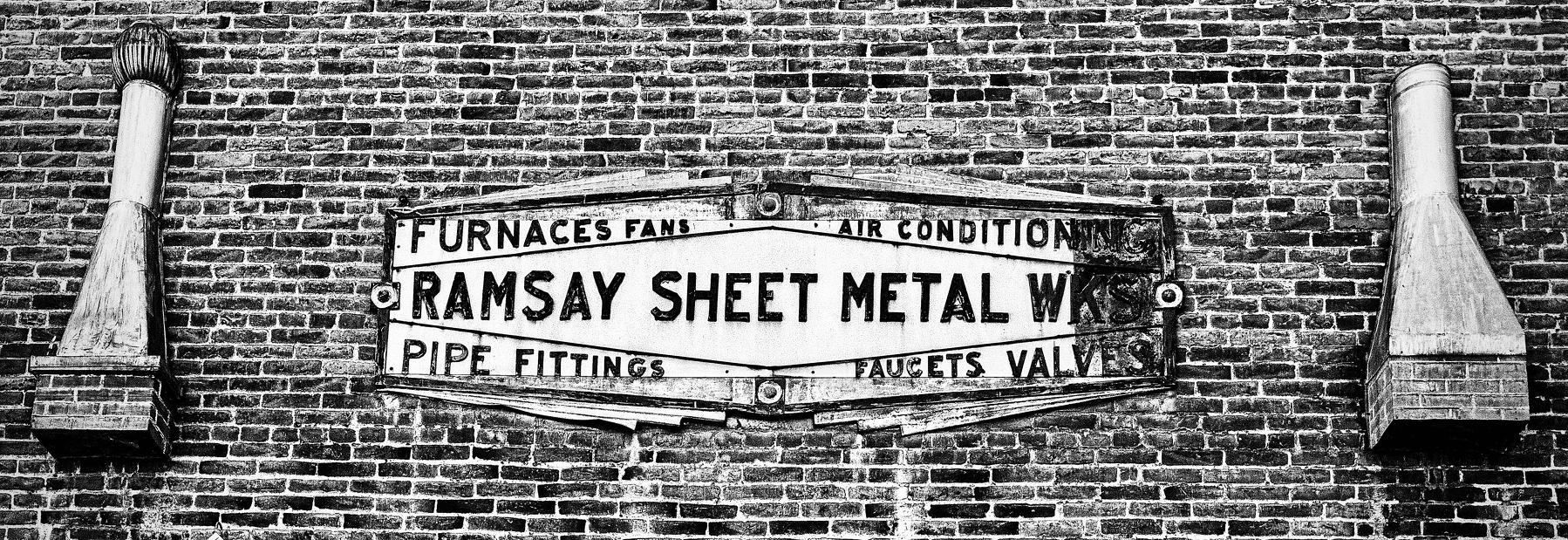 Auto-generated description: An old, weathered metal sign for Ramsay Sheet Metal Works advertising services like furnaces, fans, air conditioning, pipe fittings, faucets, and valves is mounted on a brick wall between two downspouts.