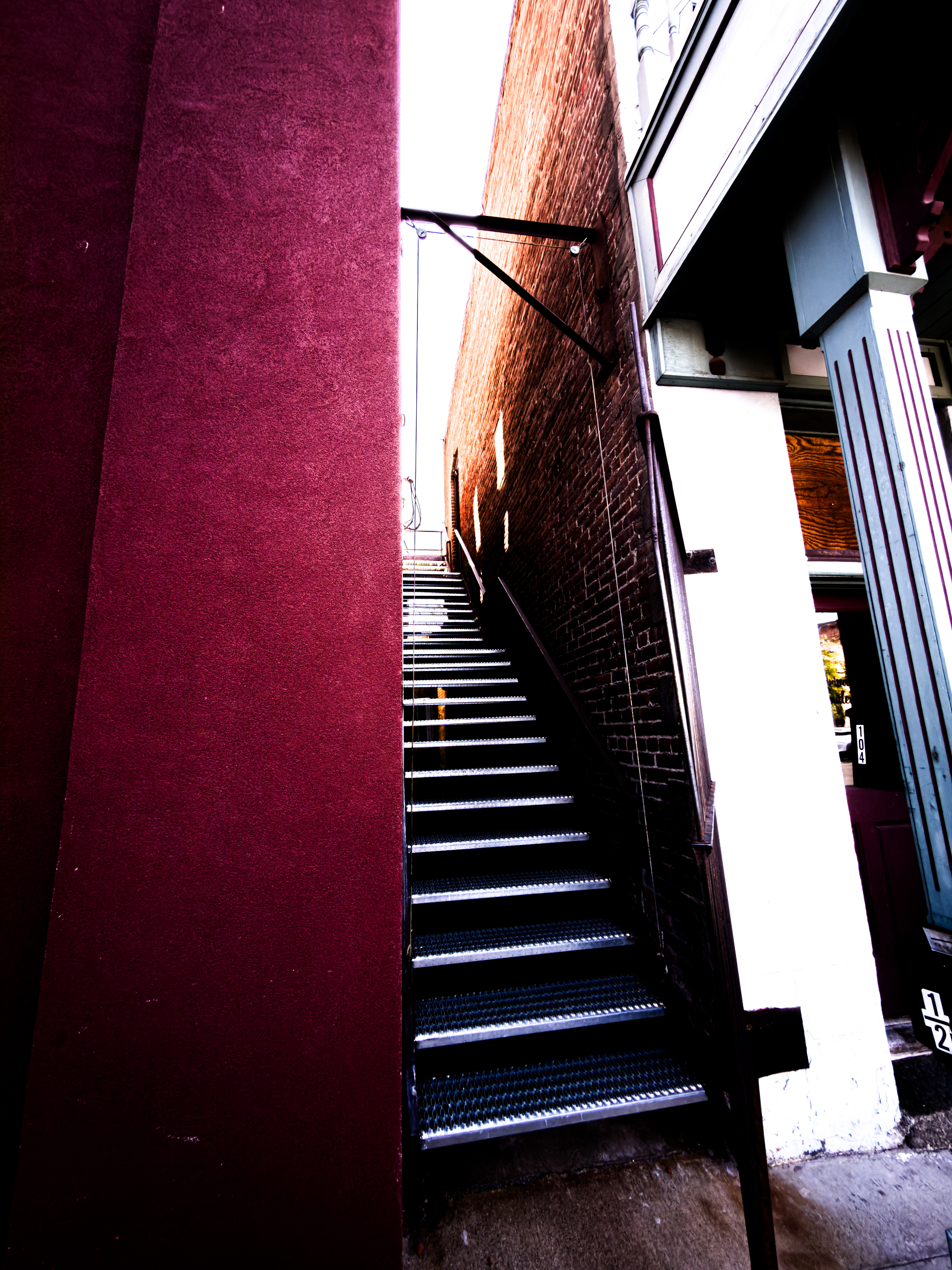Auto-generated description: A narrow metal staircase is situated between a maroon wall and a brick wall, leading upwards toward a bright opening.