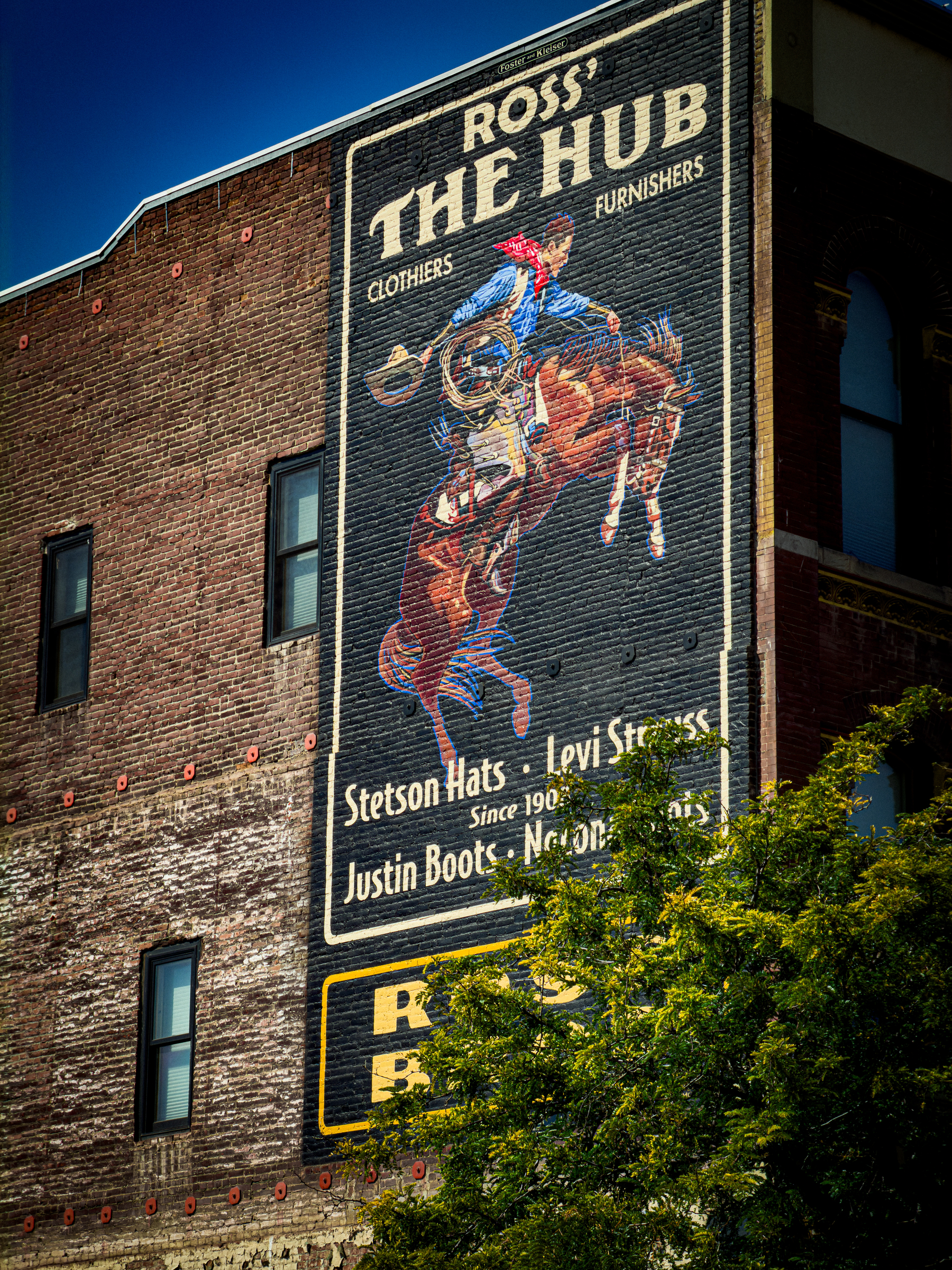 Auto-generated description: A mural on a brick building features a cowboy riding a bucking horse and advertises Ross-The Hub, a store specializing in clothes, furnishings, and brands like Stetson Hats, Levi Strauss, and Justin Boots.