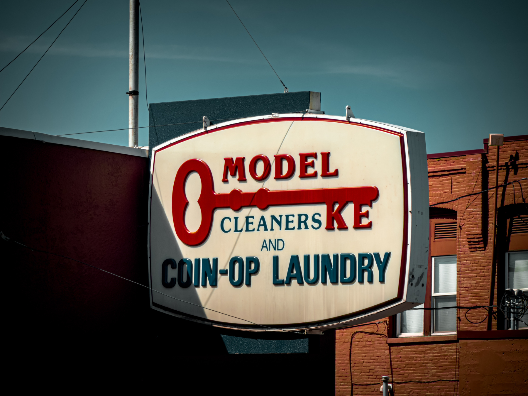 Auto-generated description: A retro-style sign for Model Key Cleaners and Coin-Op Laundry hangs against a backdrop of brick buildings and a clear sky.