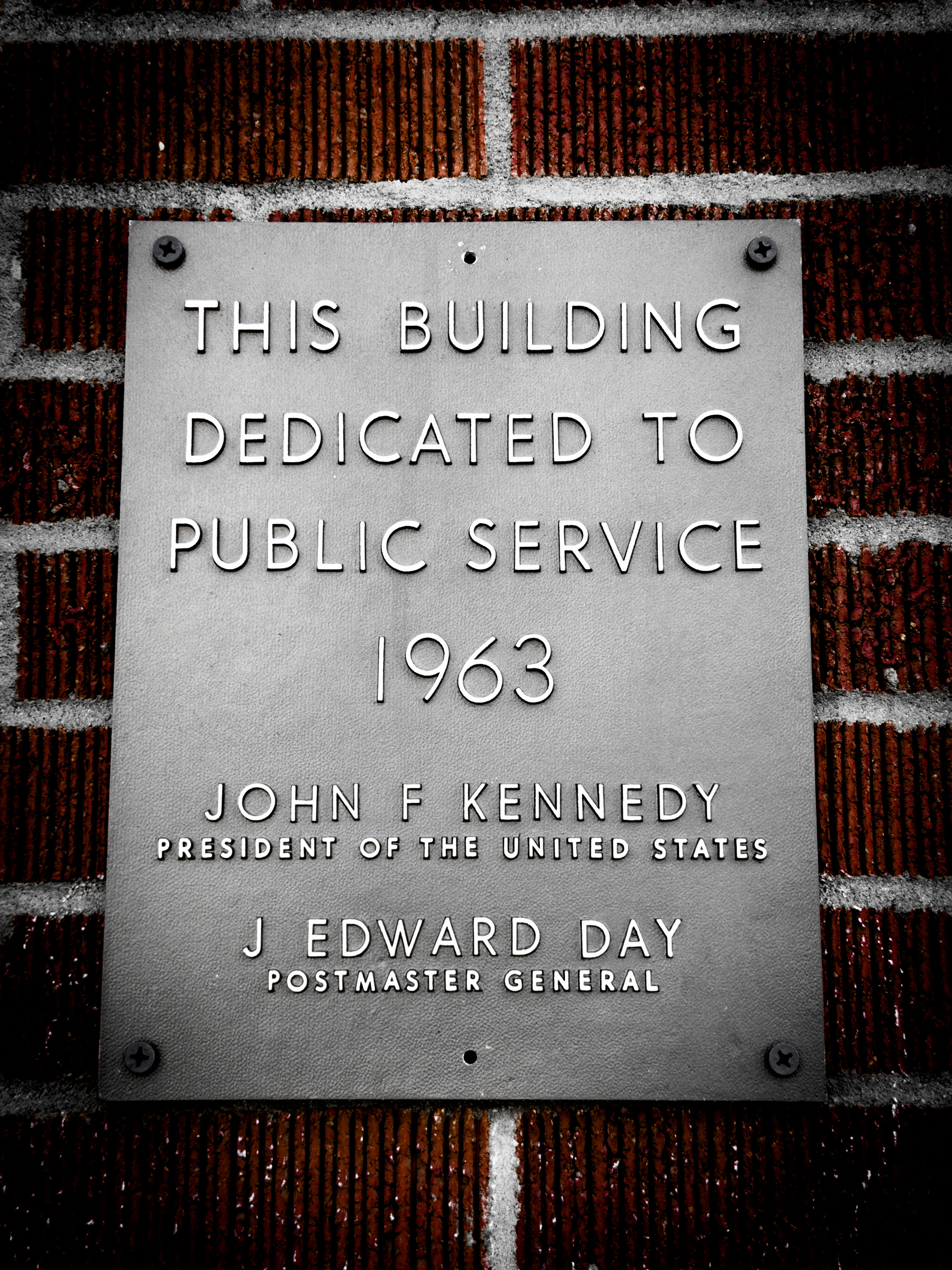 Auto-generated description: A metal plaque mounted on a brick wall dedicates the building to public service, noting the year 1963 and mentioning John F. Kennedy as President of the United States and J. Edward Day as Postmaster General.