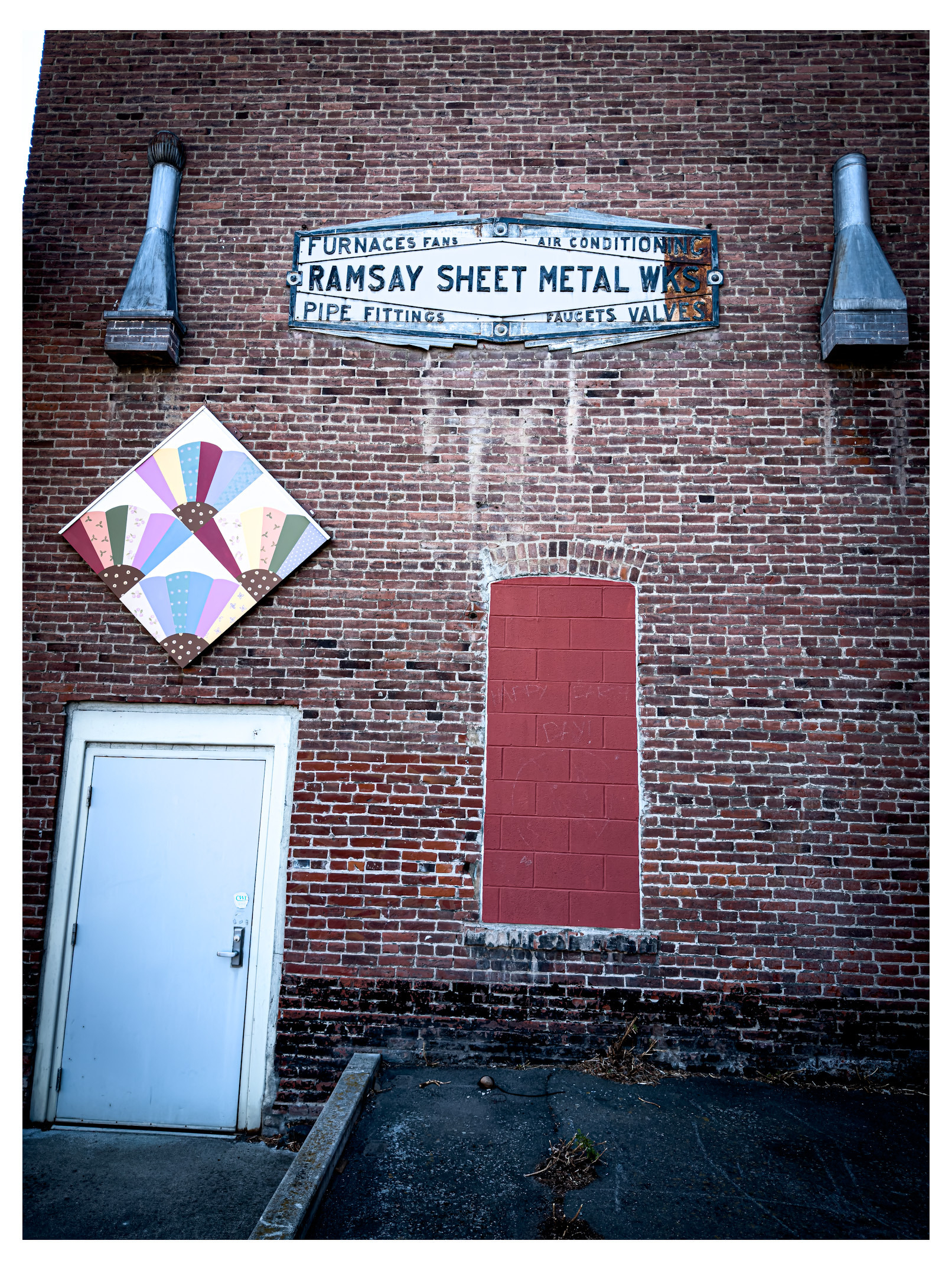 Auto-generated description: A brick wall features a faded sign for Ramsay Sheet Metal Works, two ventilation pipes, a colorful fan-shaped mural, a sealed window painted red, and a white door.