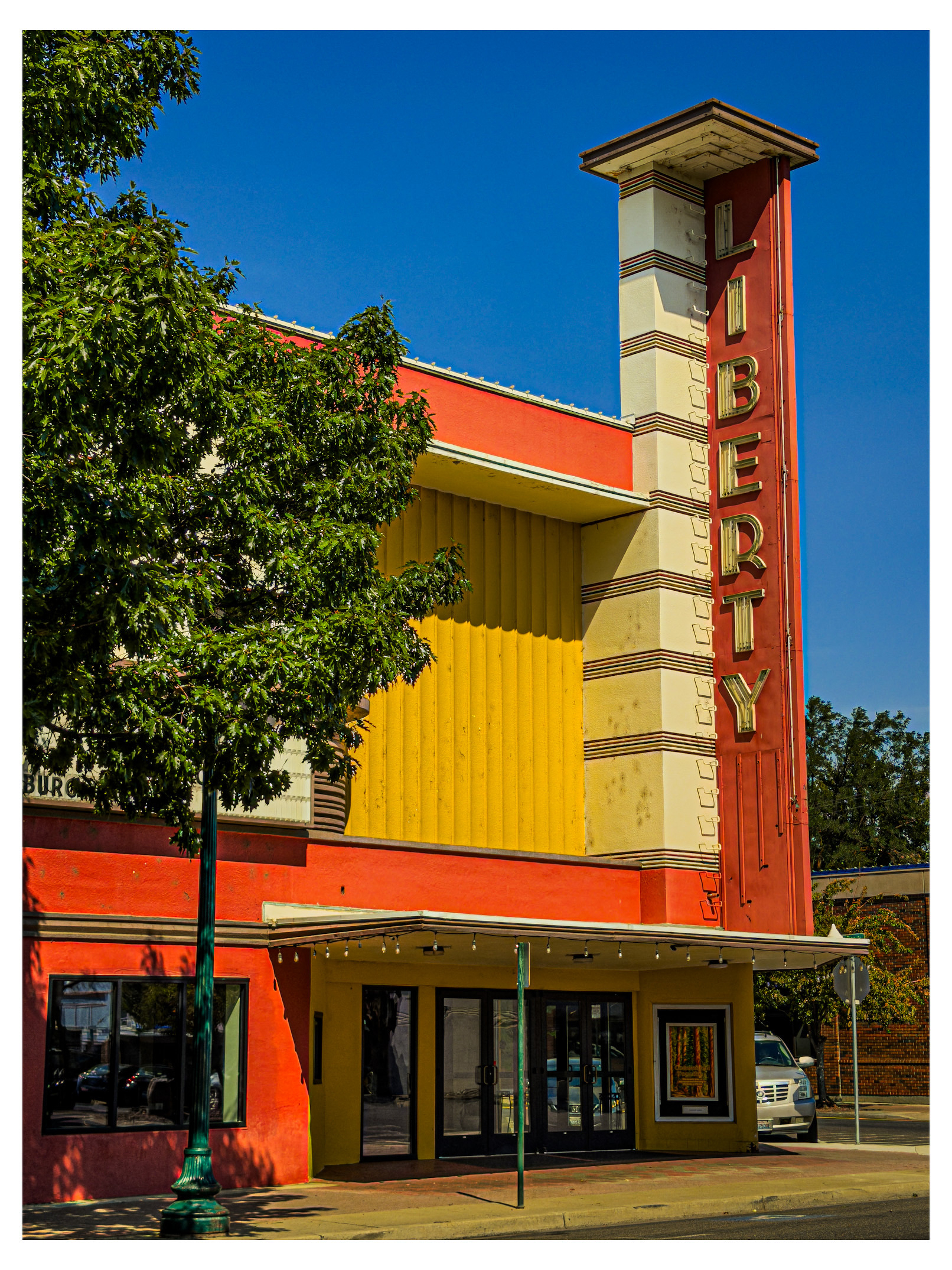 Auto-generated description: A vintage-style theater building with a tall vertical sign that reads LIBERTY stands beside a tree on a sunny day.