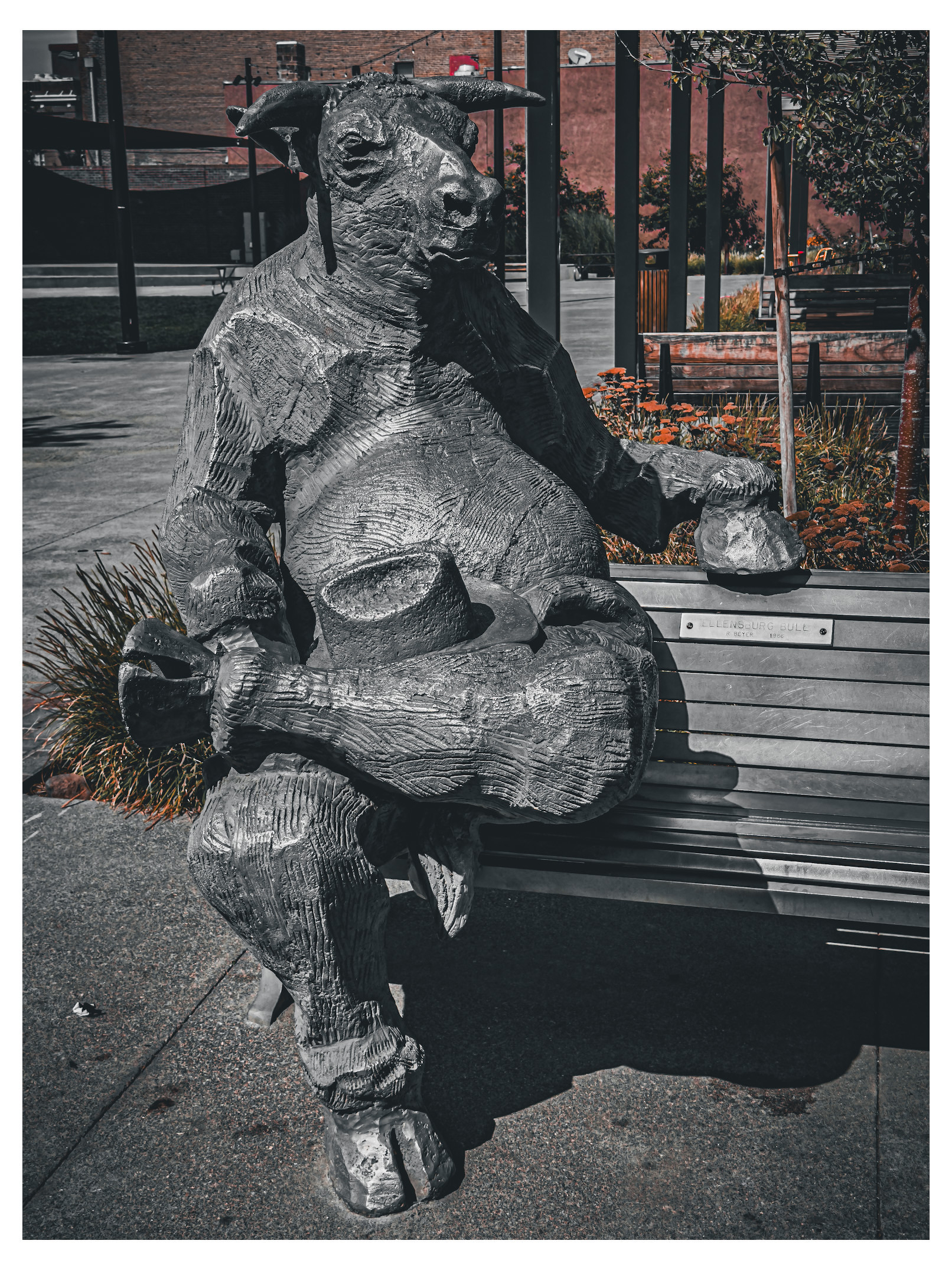 Auto-generated description: A wooden sculpture of a seated anthropomorphic bull, holding a cup and resting one leg on the opposite knee, is placed on a bench in an outdoor setting.