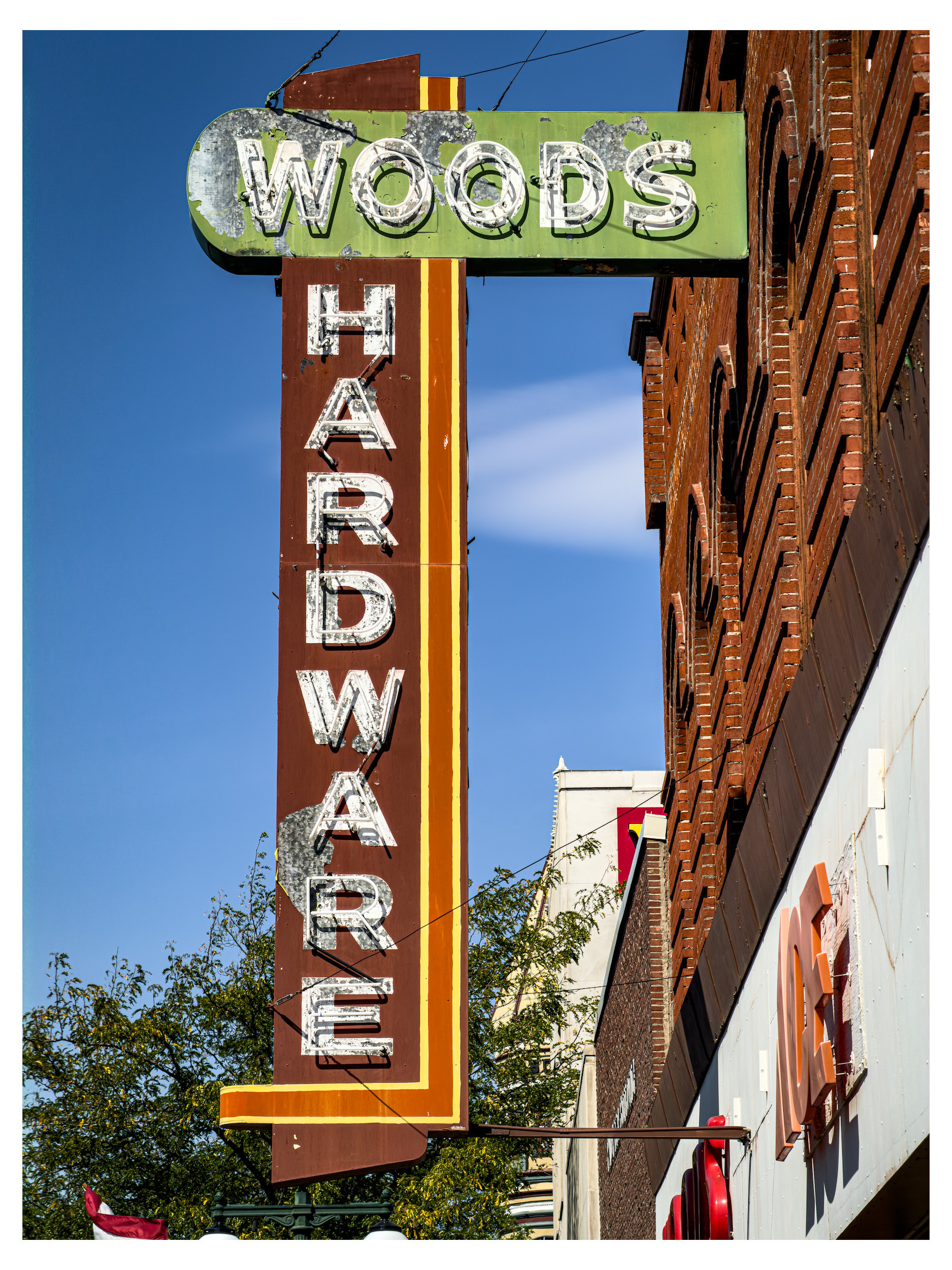 Auto-generated description: A vintage vertical sign reads Woods Hardware and is mounted on the exterior brick wall of a building against a clear blue sky.
