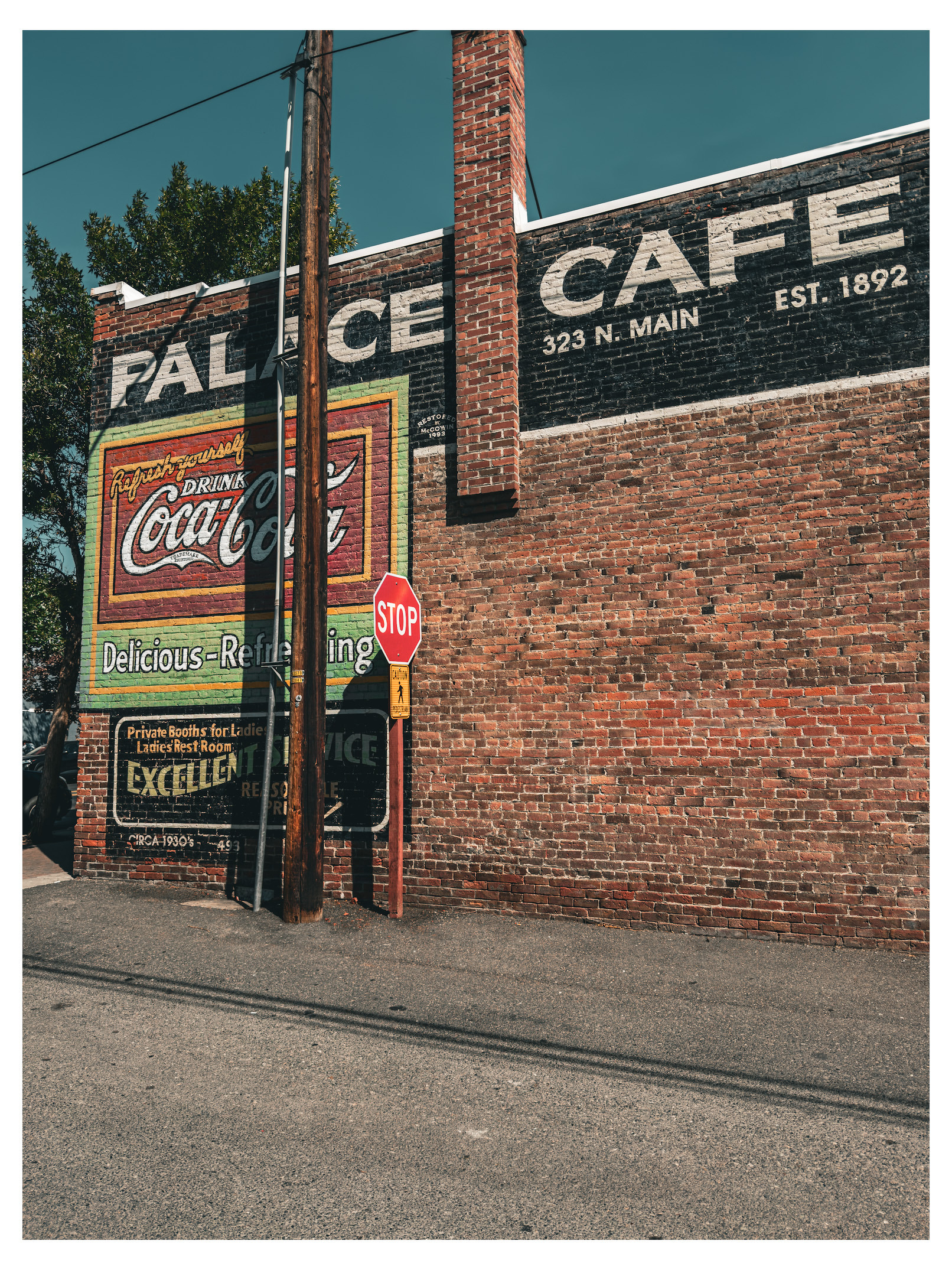 Auto-generated description: A brick building features vintage painted advertisements for Coca-Cola and the Palace Cafe at 323 N. Main, alongside a stop sign and utility poles.