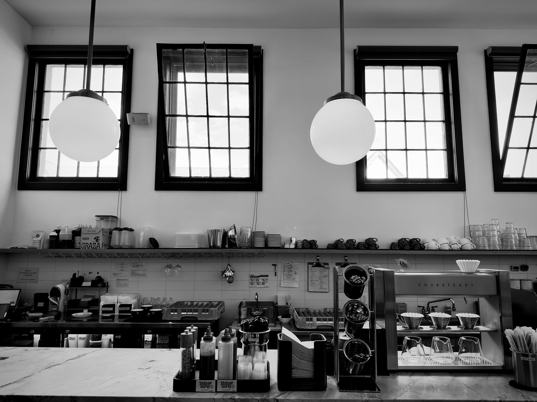 Auto-generated description: A minimalist coffee shop counter features various coffee-making equipment, condiments, and three large pendant lights hanging below tall, gridded windows.