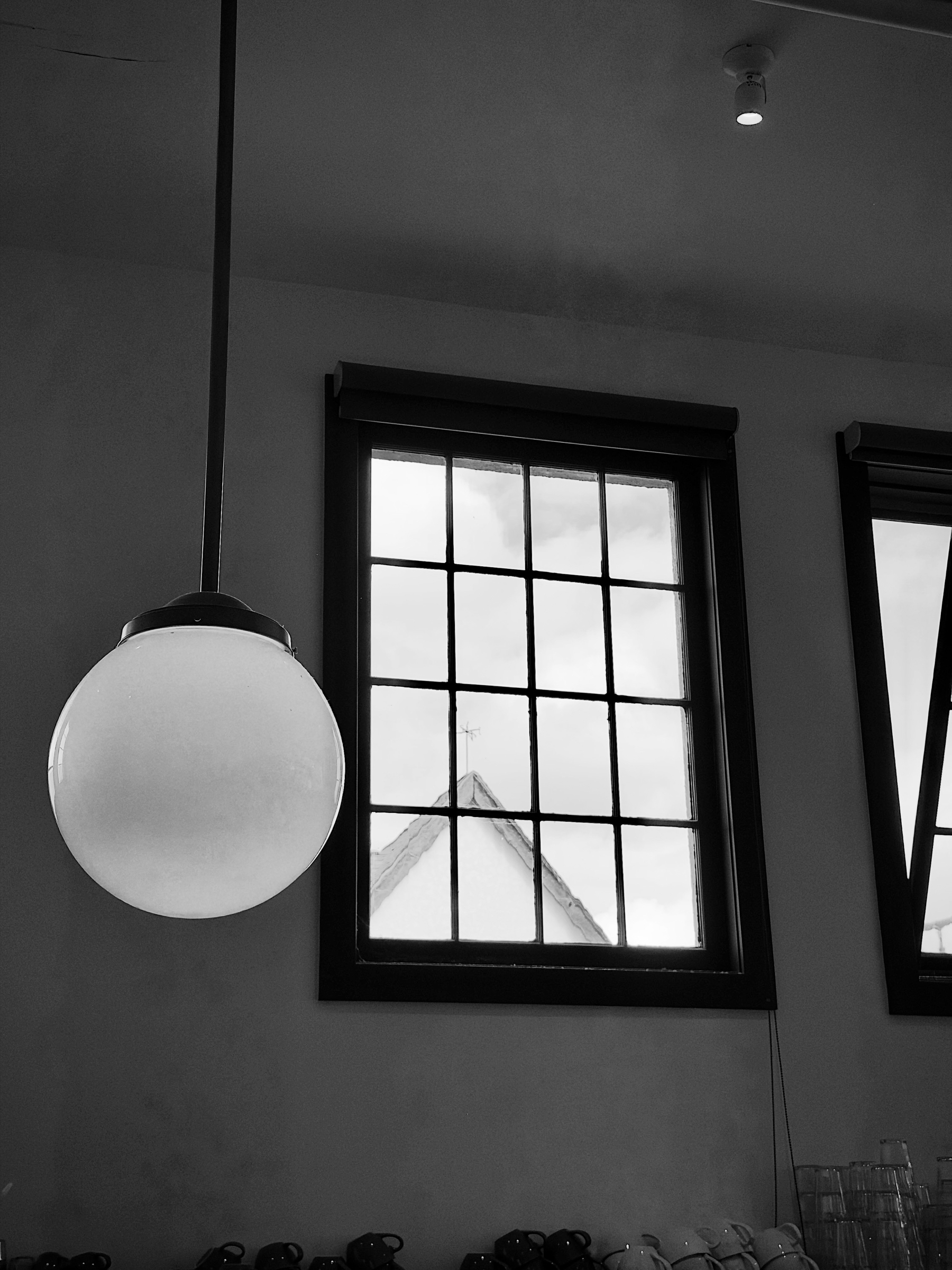 Auto-generated description: A round pendant light hangs near a window with a grid pattern, through which the peak of a roof and cloudy sky are visible.