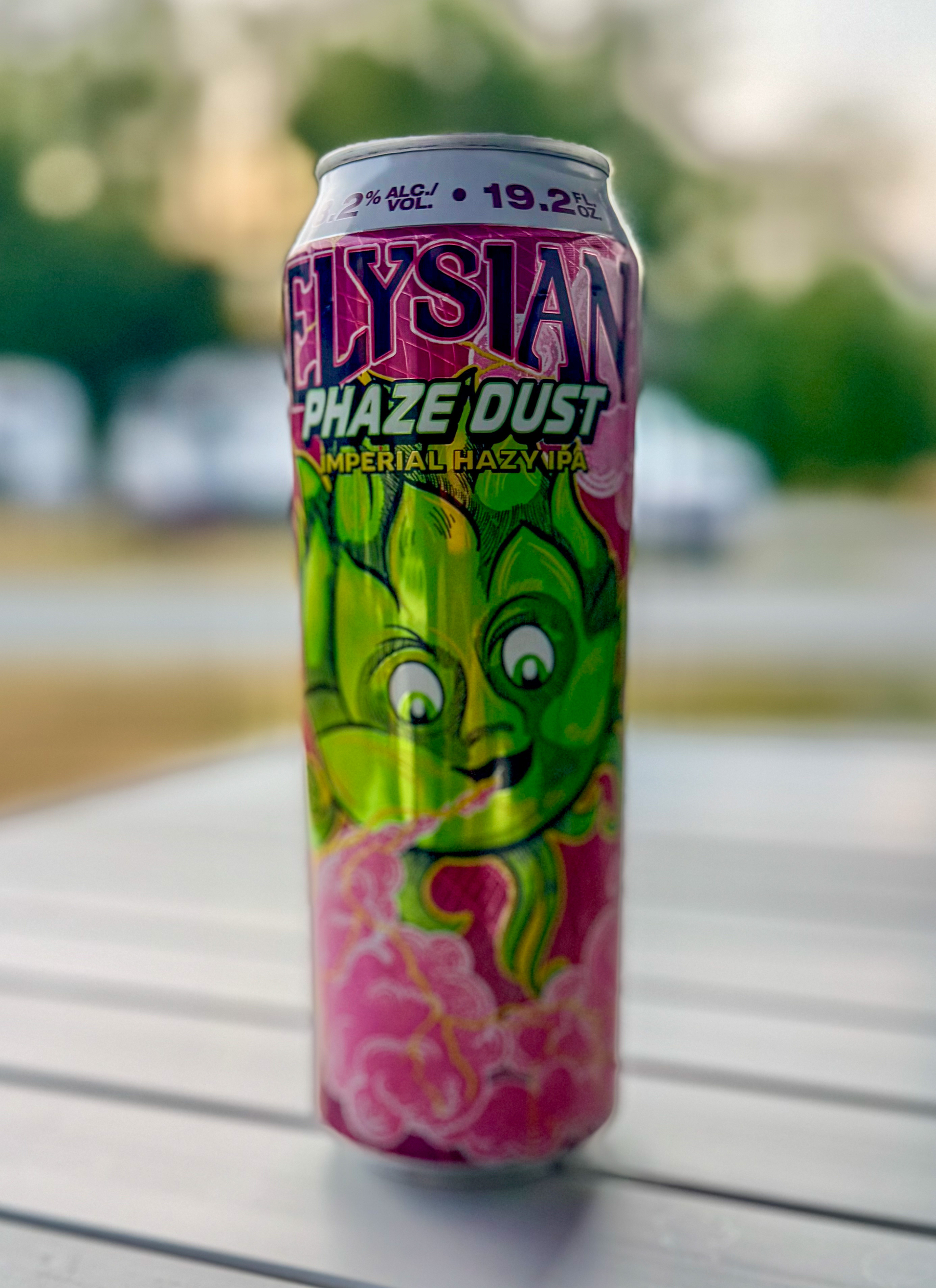 Auto-generated description: A colorful can of Elysian Phaze Dust Imperial Hazy IPA, featuring a green hop character and displaying an alcohol content of 9.2% and a volume of 19.2 fl oz, is placed on a white surface with a blurred outdoor background.