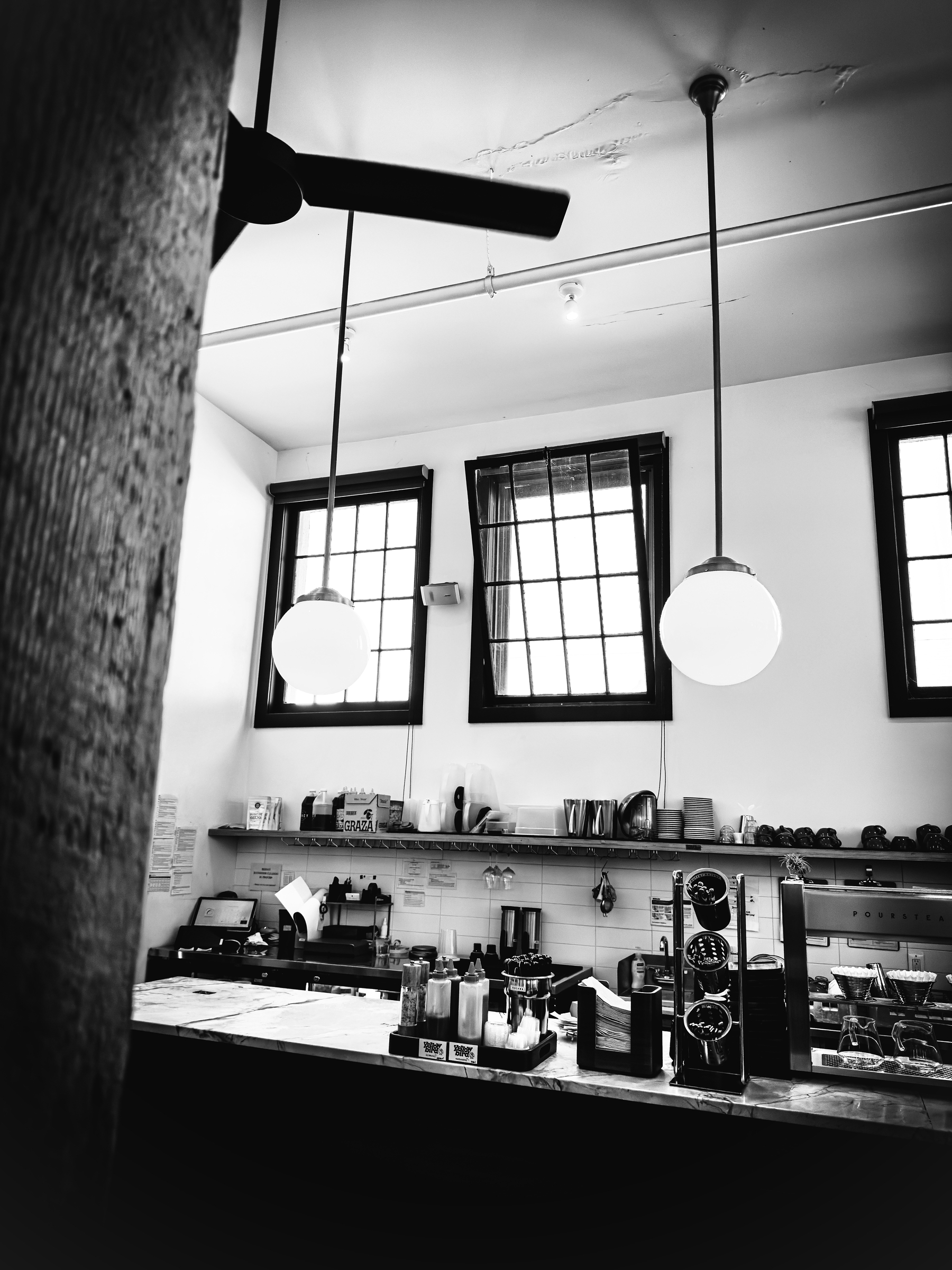 Auto-generated description: A black and white photo shows a coffee shop counter with hanging globe lights, windows, and various coffee-making equipment.