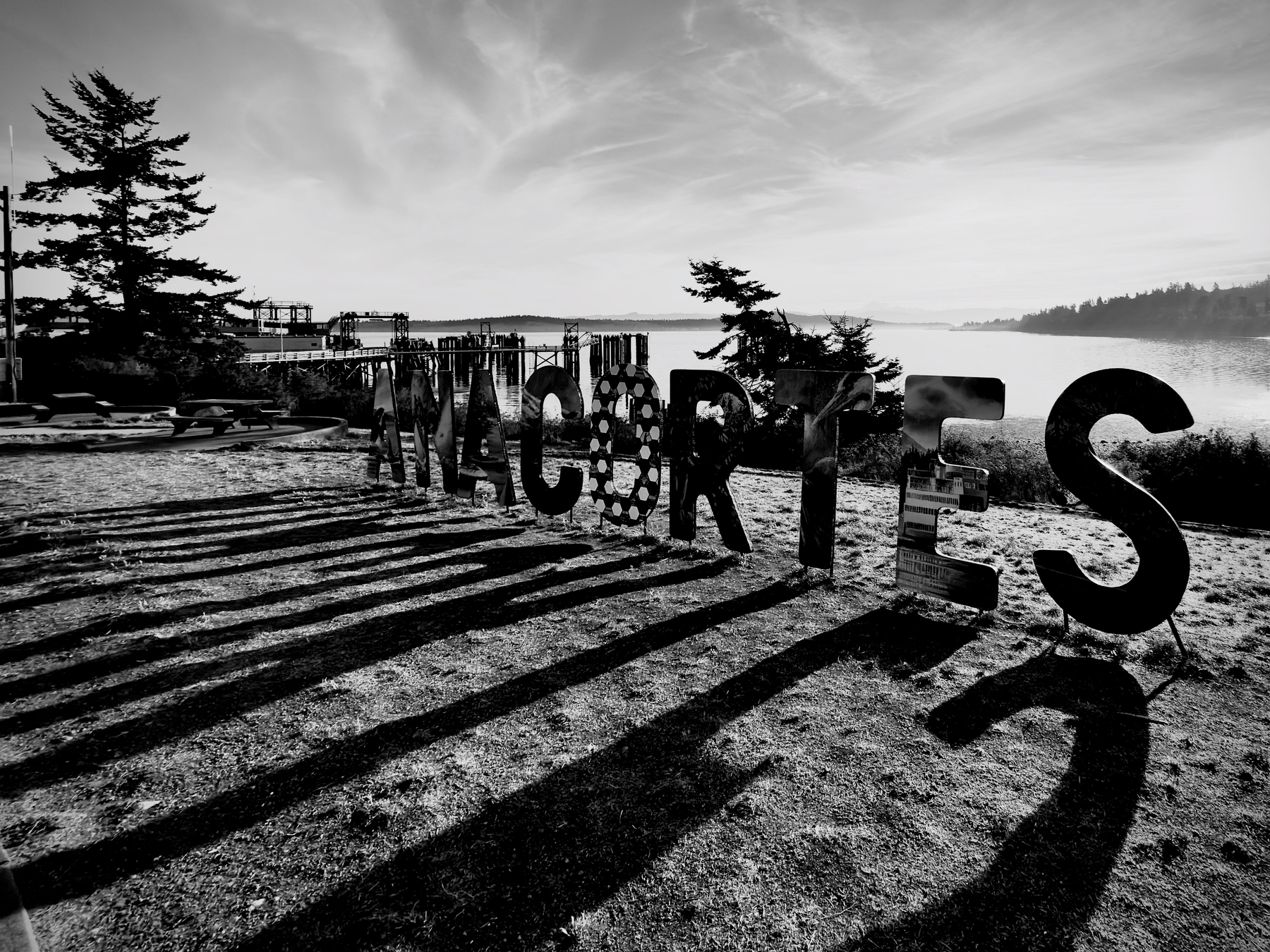 Auto-generated description: Large silhouetted letters spelling WALCARTES stand on a beach near a dock, casting long shadows in the light of the setting or rising sun.
