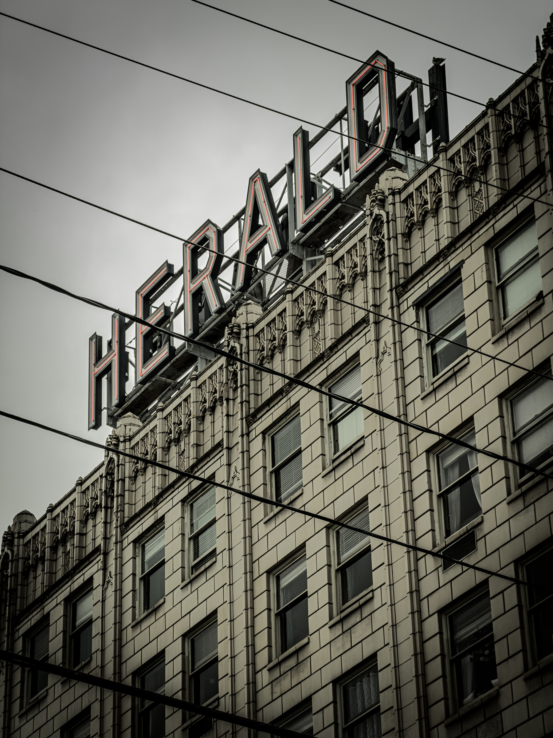 Auto-generated description: A tall, ornate building features a large HERALD sign on its roof with power lines crossing in front against a cloudy sky.