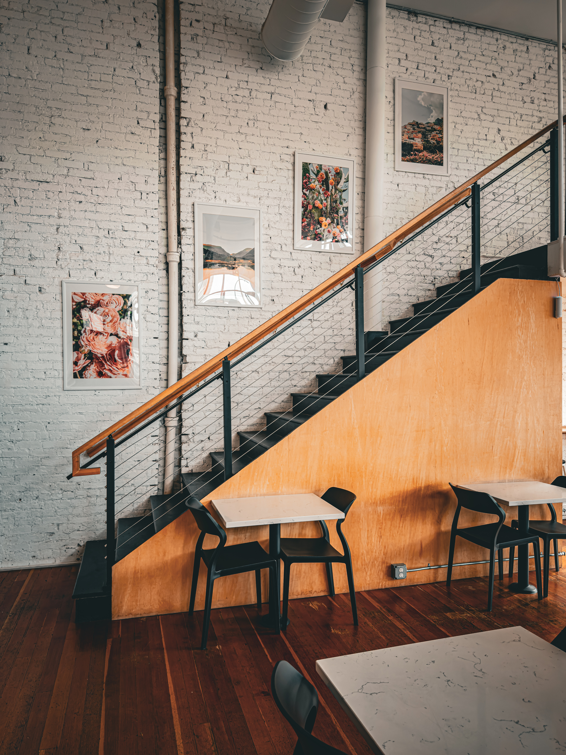 Auto-generated description: A cozy cafe interior features wooden floors, a staircase with a black railing and wooden paneling, and framed floral and nature artwork on a white brick wall.