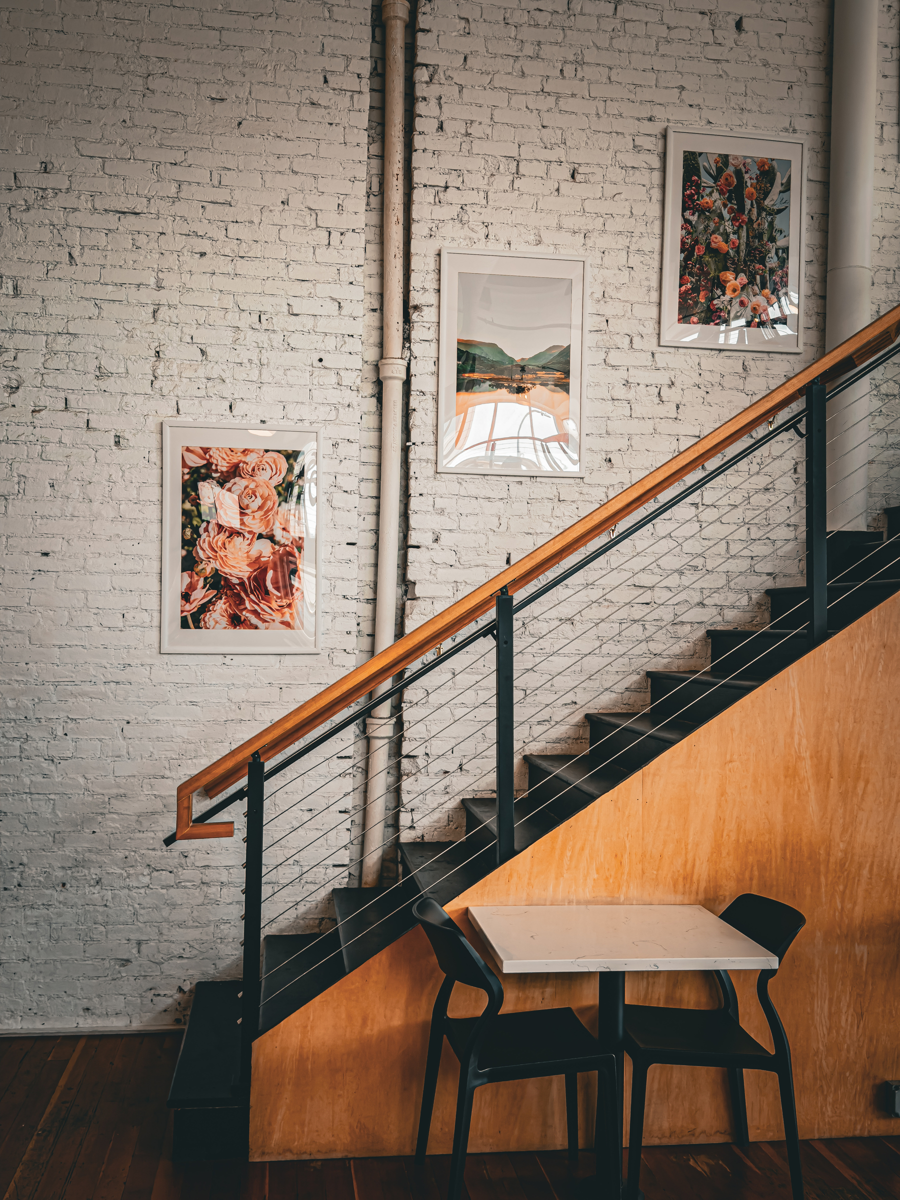 Auto-generated description: A small table with two black chairs is placed under a staircase decorated with three framed pictures on a white brick wall.