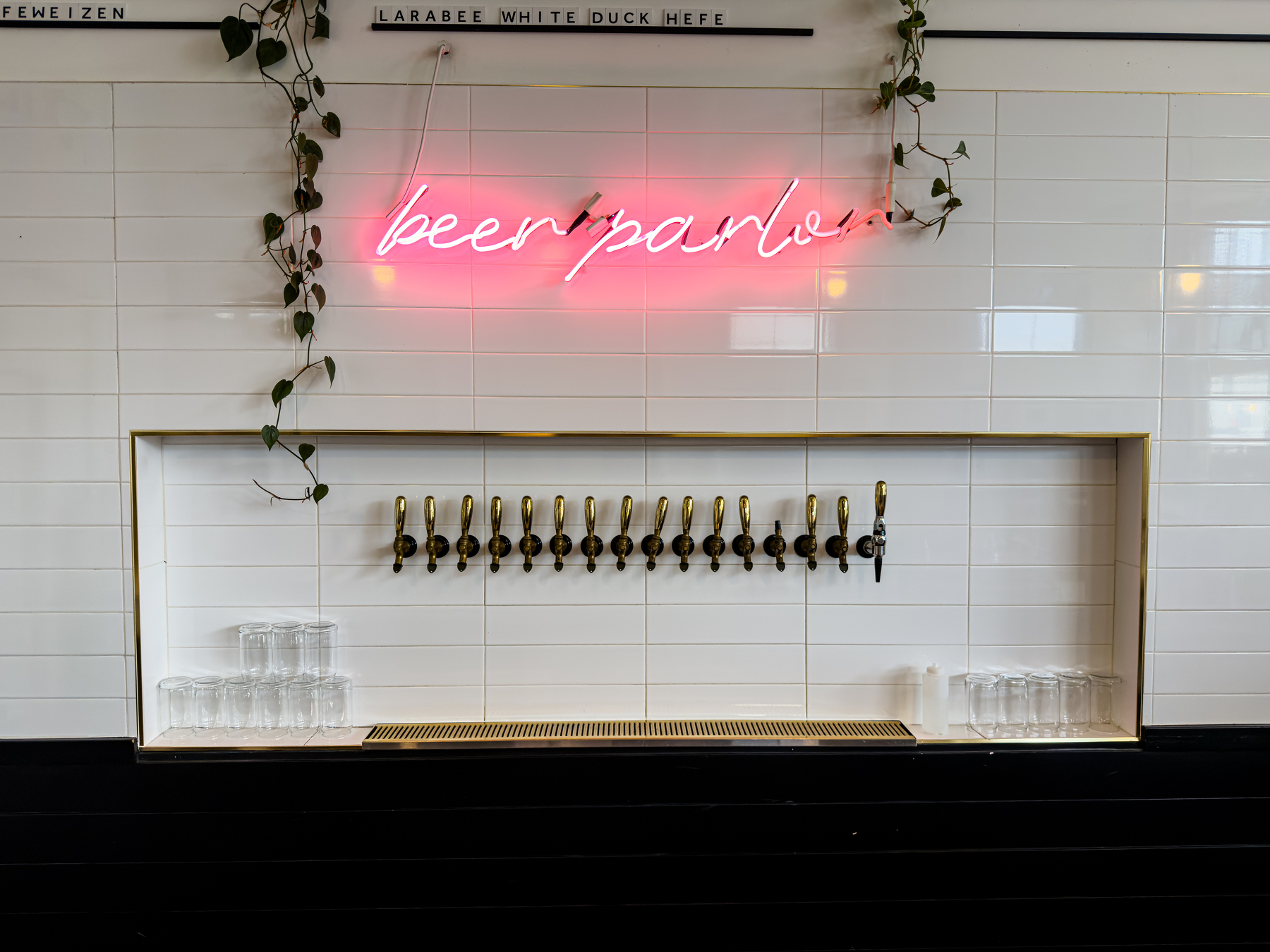 Auto-generated description: A row of beer taps is mounted on a white tiled wall with a pink neon sign reading beer parlor and vines hanging above.