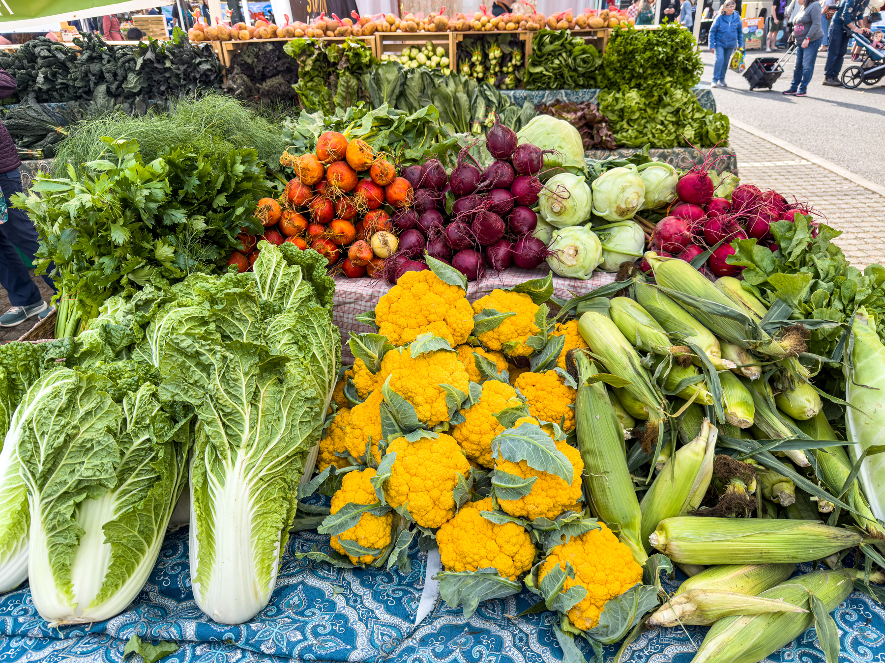 Auto-generated description: A colorful display of fresh vegetables including orange cauliflower, corn, leafy greens, beets, and kohlrabi is arranged on a market stall.