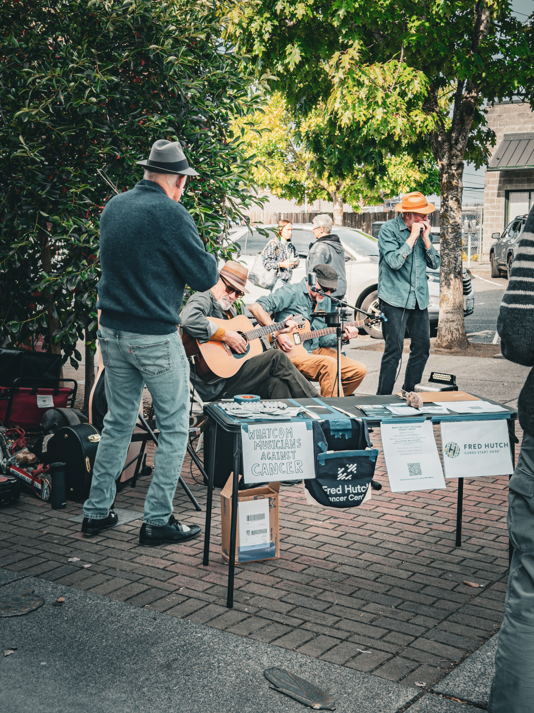 Auto-generated description: A group of musicians is performing outdoors near a table with signs supporting the Fred Hutch Cancer Center and a cause related to cancer awareness.