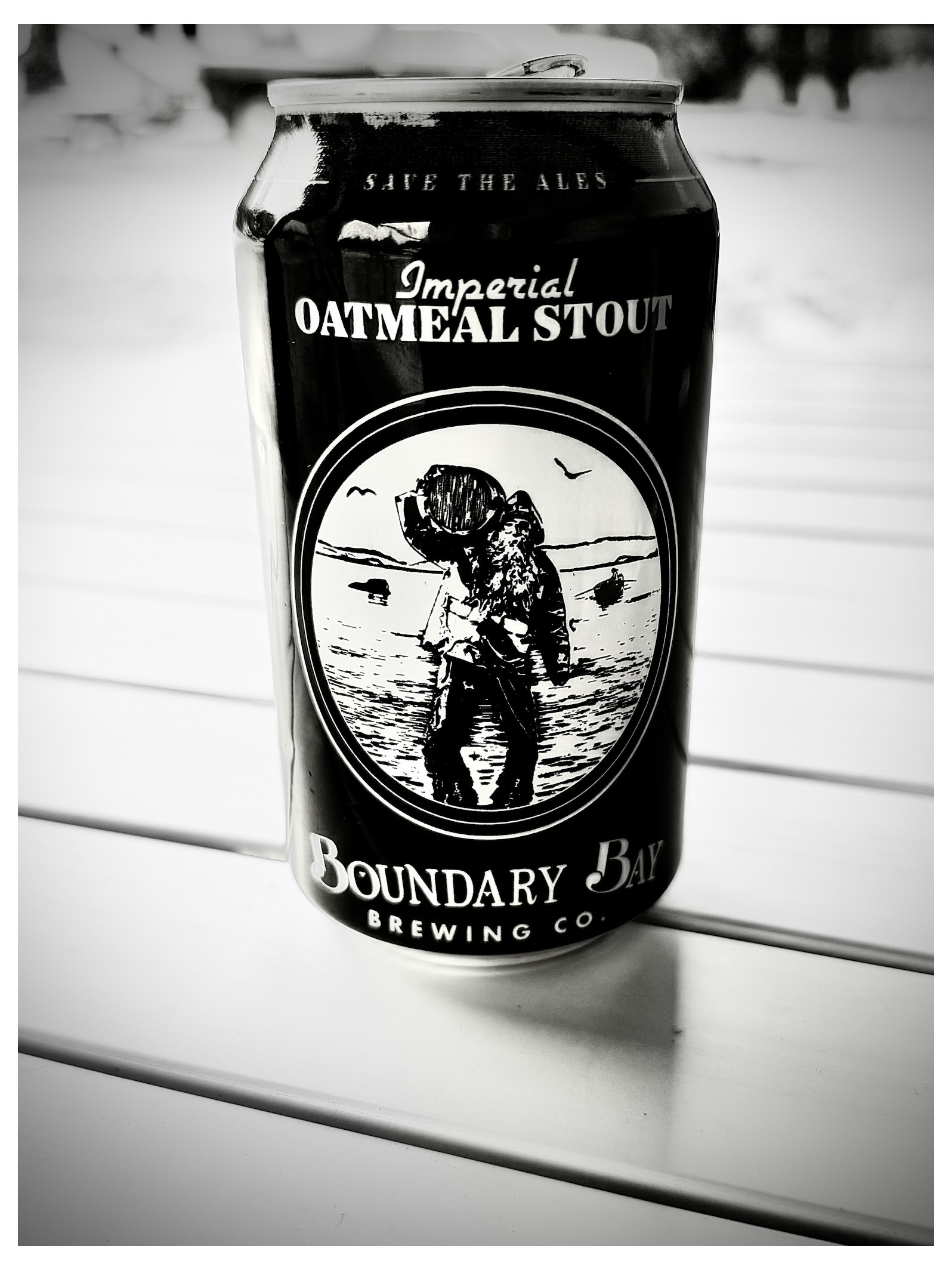 Auto-generated description: A black can of Boundary Bay Brewing Co.'s Imperial Oatmeal Stout with a vintage-style illustration of a man carrying a keg is placed on a white surface.