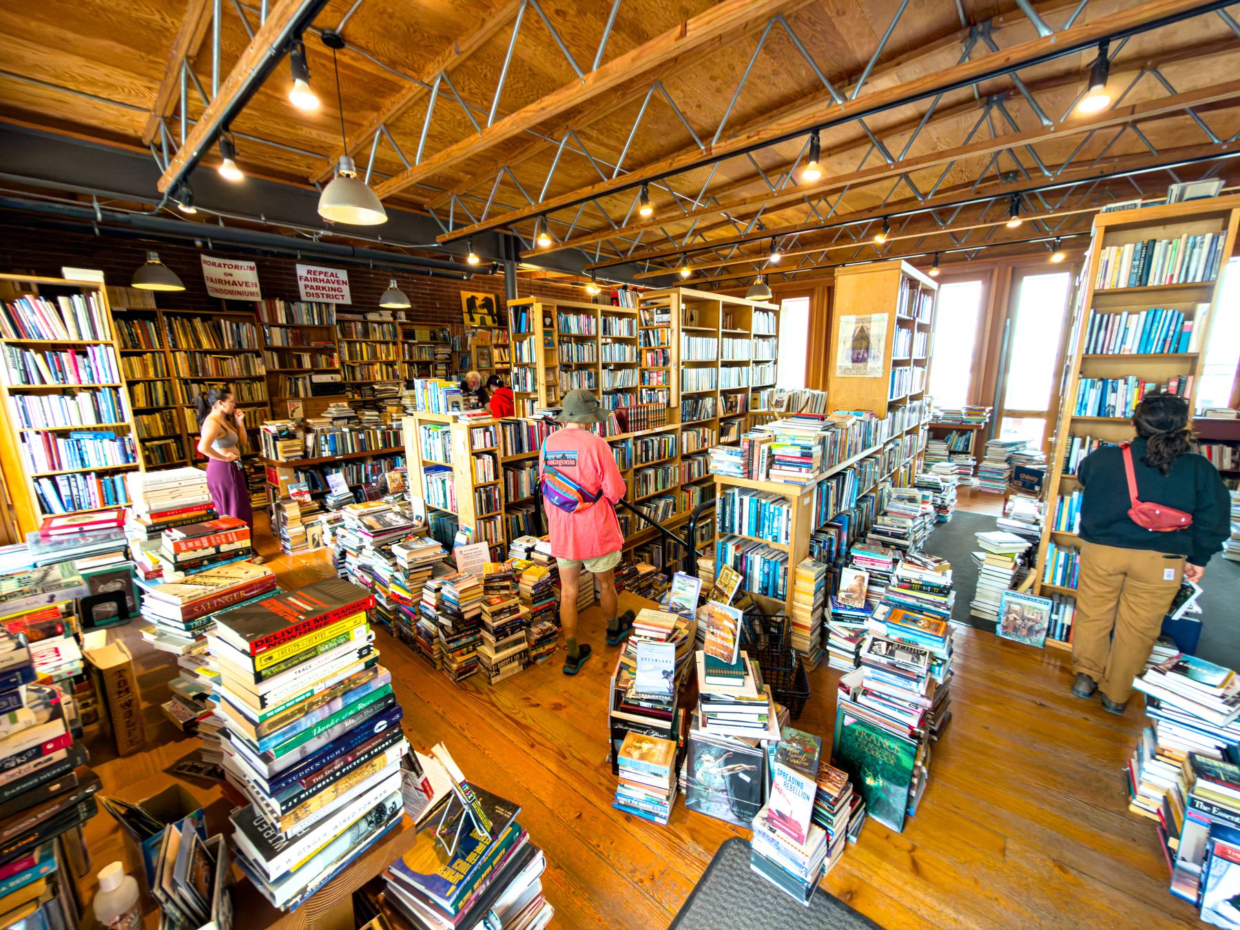 Auto-generated description: A bustling bookstore with tall wooden shelves and stacks of books on the floor, filled with several people browsing.
