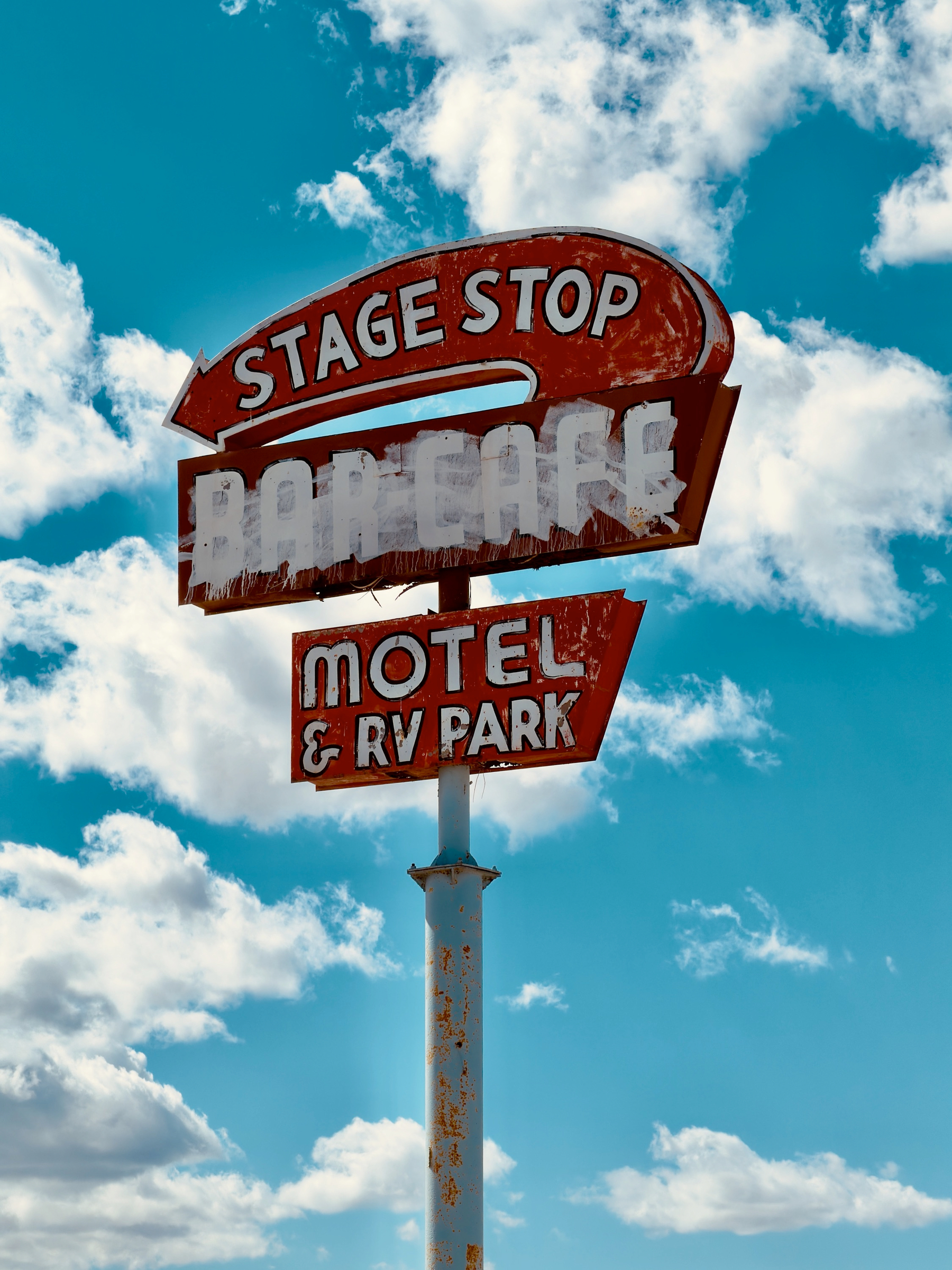 Auto-generated description: A vintage red sign for a motel and RV park with the words Stage Stop against a blue sky with scattered clouds.