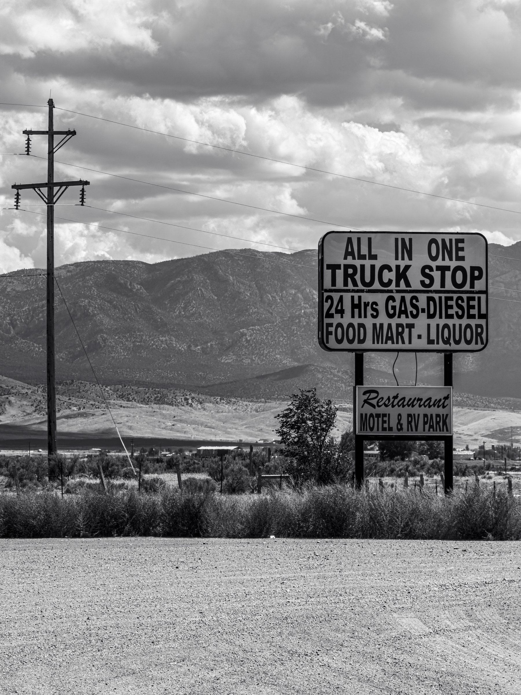 Auto-generated description: A roadside sign advertises an all-in-one truck stop offering 24-hour gas, diesel, food mart, liquor, restaurant, motel, and RV park services against a mountainous backdrop.